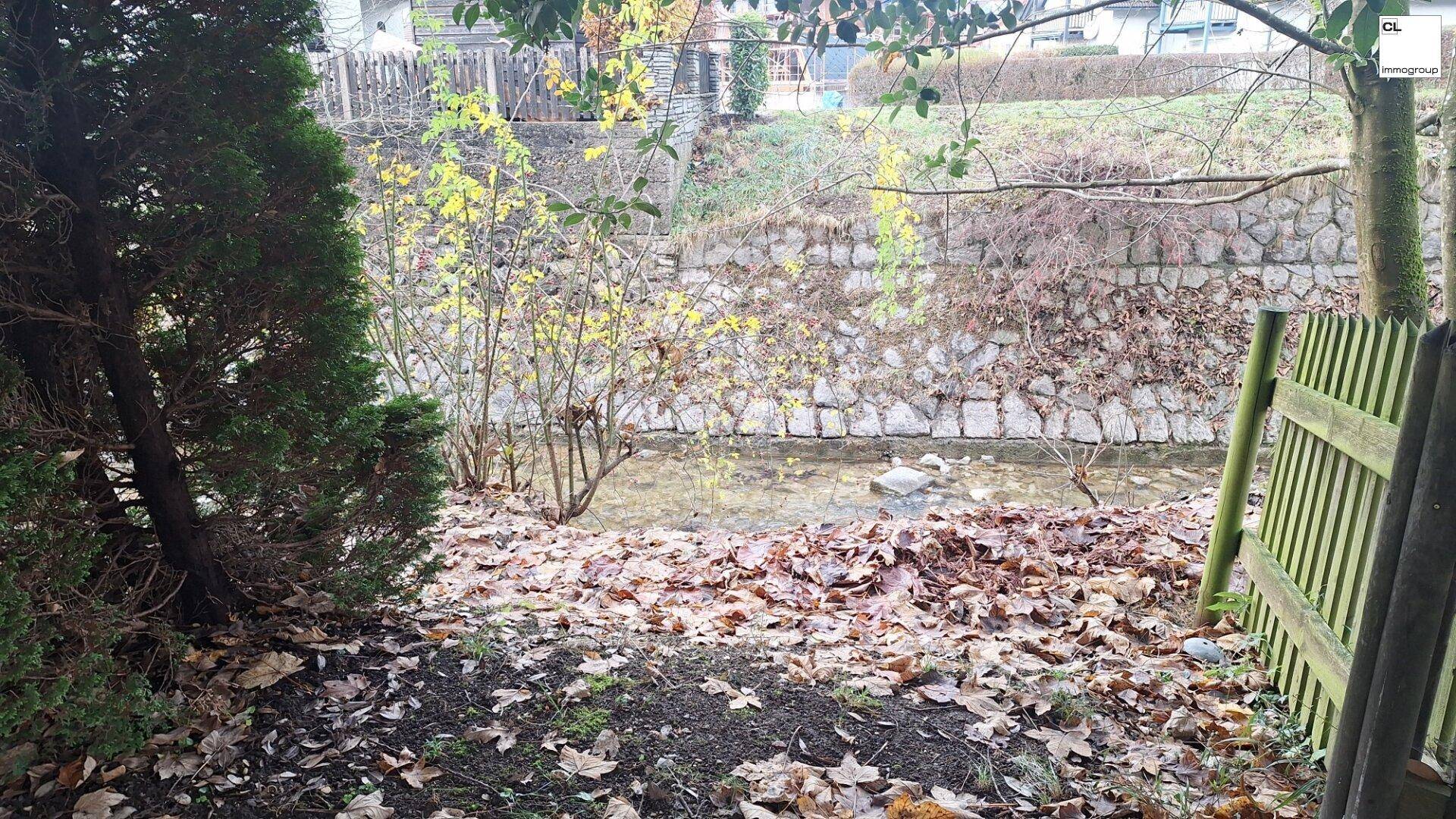 Kleiner Bach oder Kanal mit Steinmauer und üppiger Vegetation, umgeben von Bäumen und Laub.
