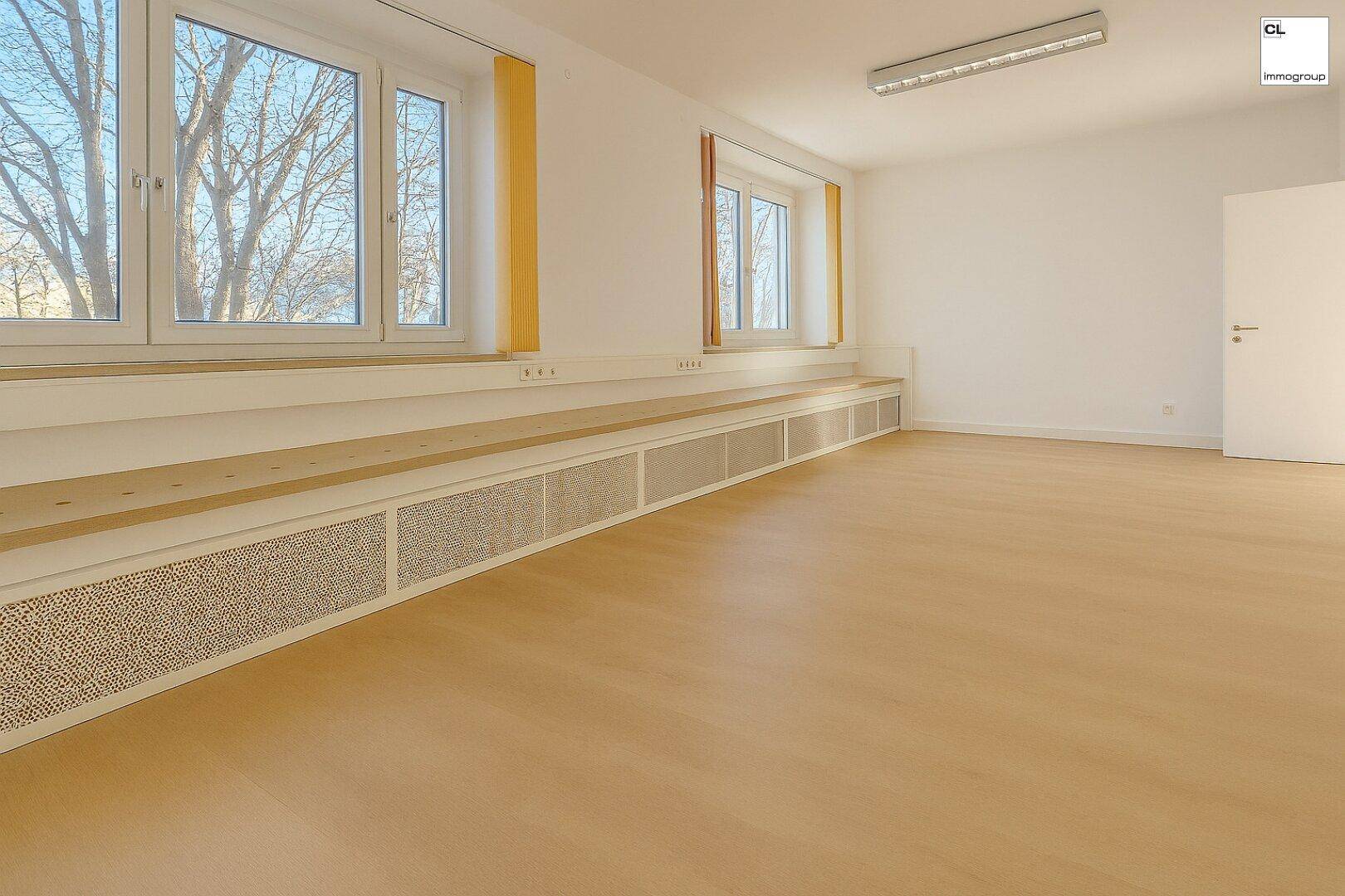 Heller, leerer Raum mit großen Fenstern, Holzboden und einer langen Sitzbank entlang der Wand.