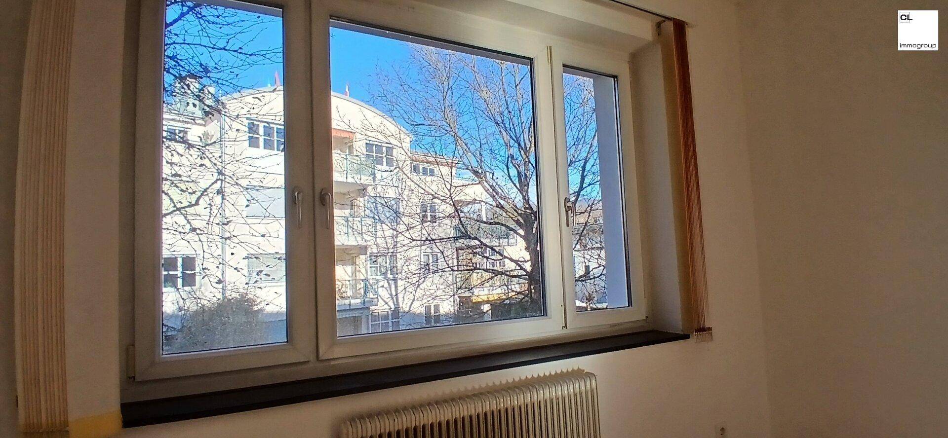 Großes Fenster in einem hellen Raum mit Blick auf andere Gebäude und Bäume.