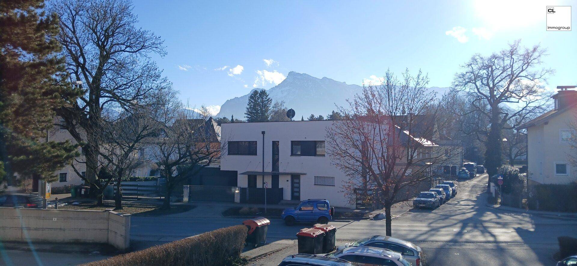 Panoramablick auf eine Wohngegend mit Bäumen, Häusern und Bergen im Hintergrund unter blauem Himmel.