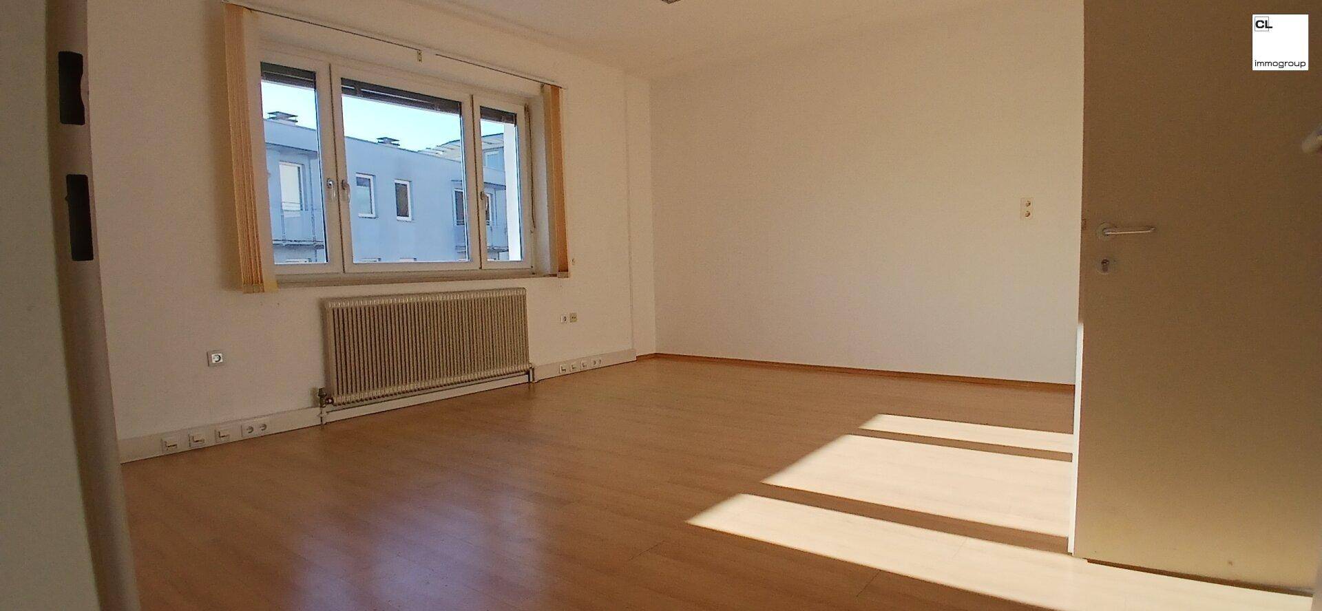 Heller, leerer Raum mit Holzboden, großem Fenster und Heizkörper unter dem Fenster.