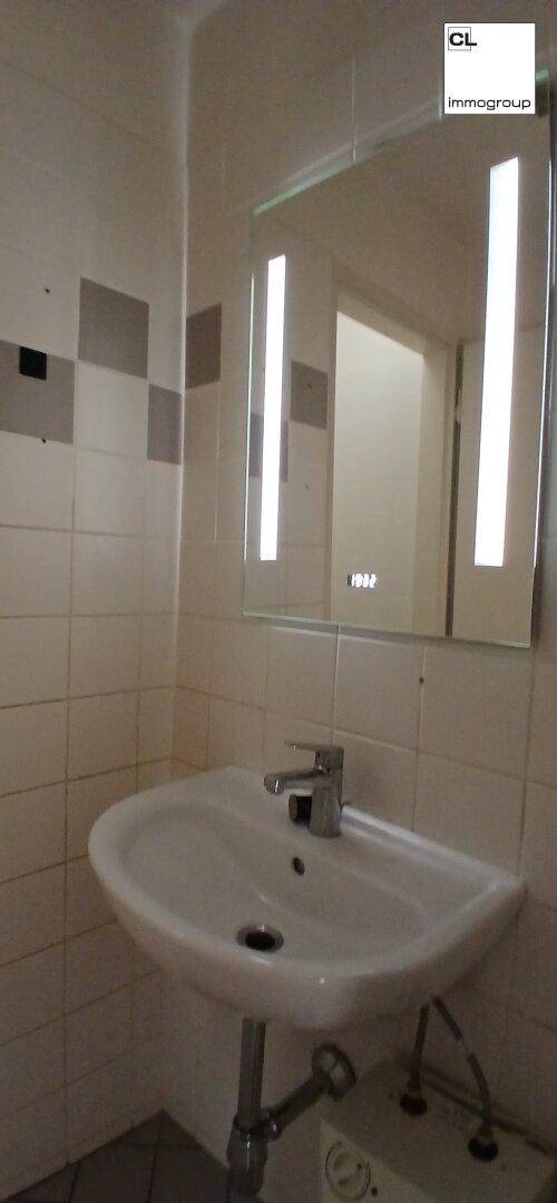 Kleines Badezimmer mit Waschbecken, beleuchtetem Spiegel und weiß-grauen Fliesen an den Wänden.