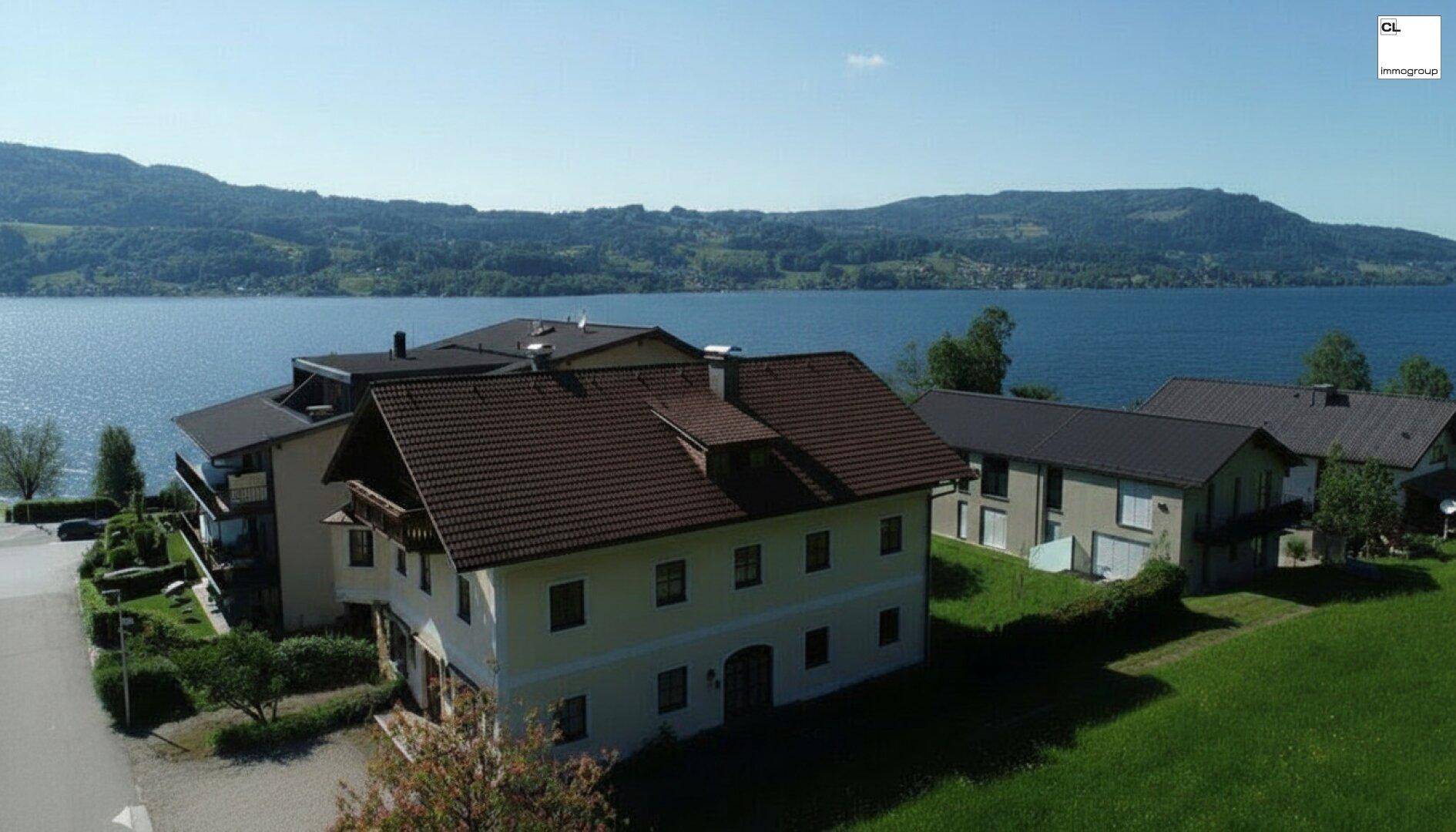 Luftaufnahme eines Anwesens am Seeufer mit umliegenden Häusern und Bergen im Hintergrund.