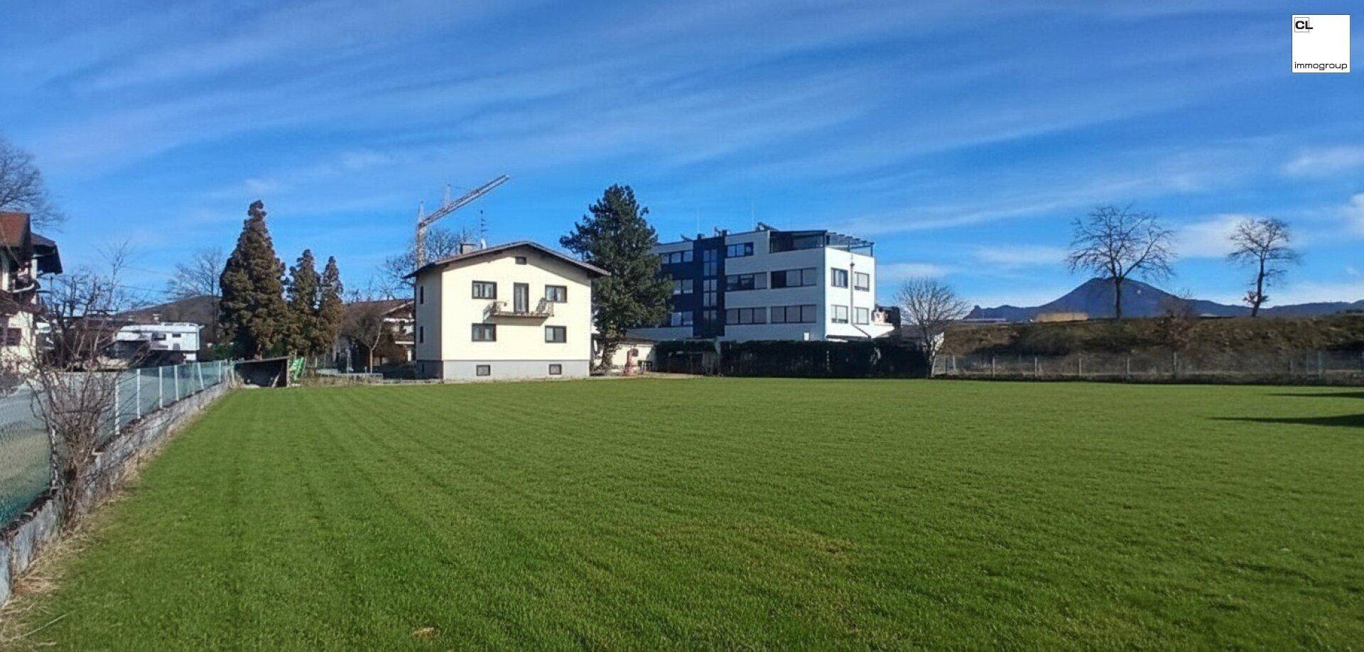 Weitläufige grüne Wiese mit Wohngebäuden und einem Neubau im Hintergrund unter klarem Himmel.