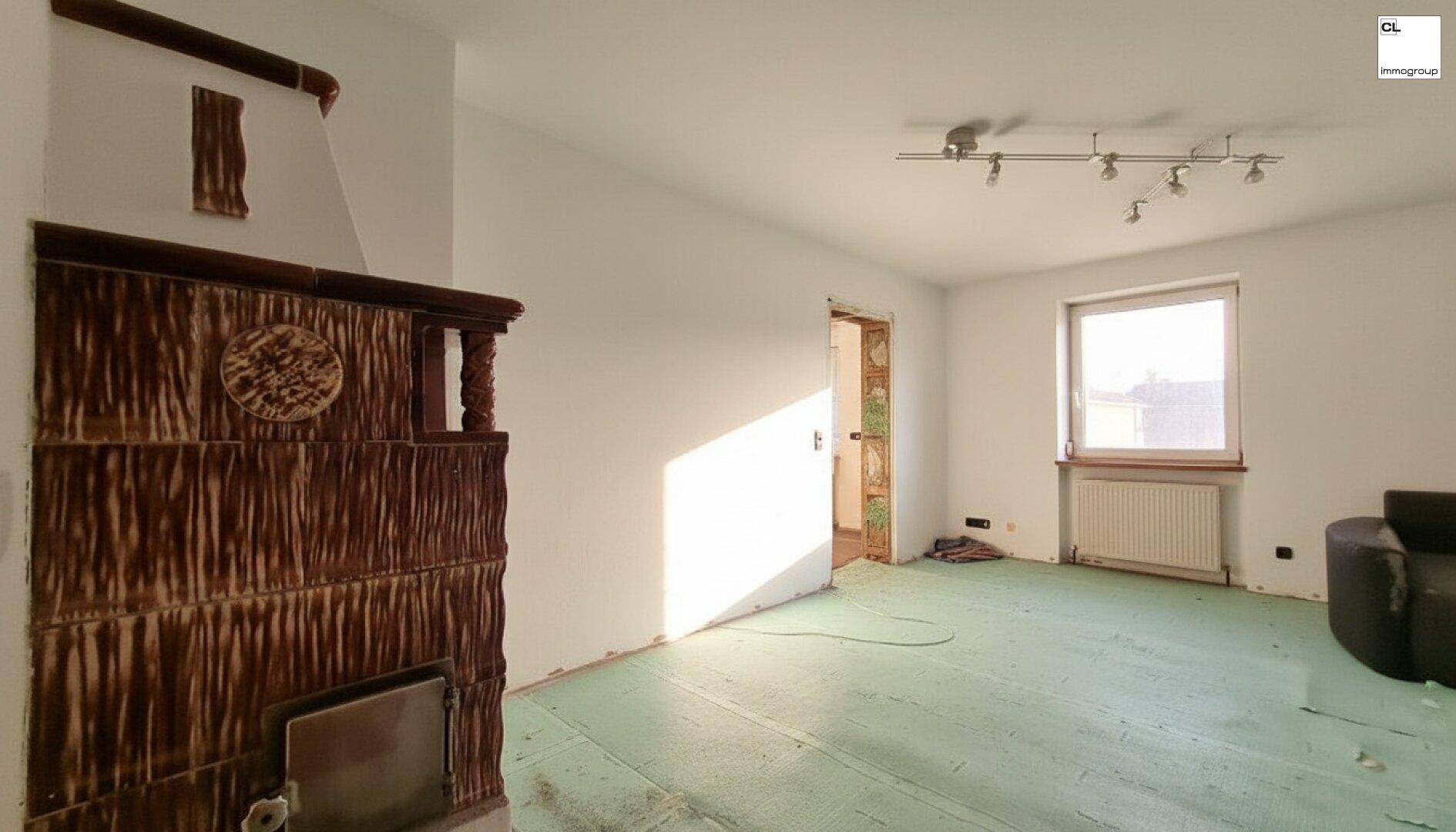 Wohnraum mit traditionellem Kachelofen, hellem Fenster und vorbereitetem Boden für neuen Belag.
