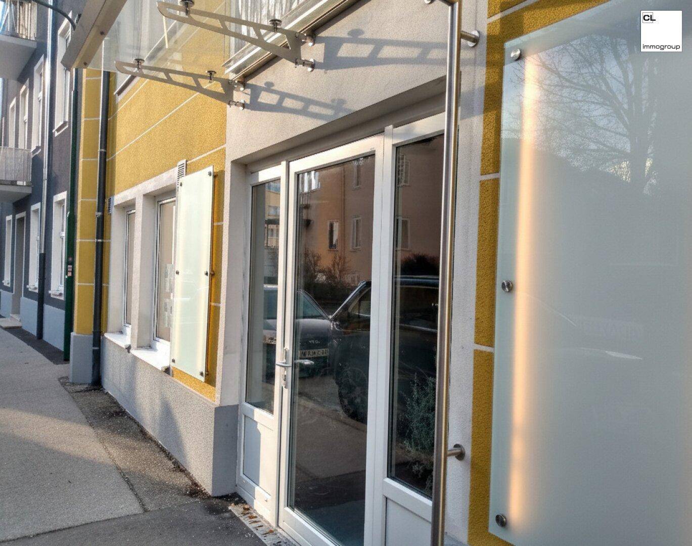 Eingangsbereich des Gebäudes mit gelber Fassade, großen Fenstern und schützendem Glasvordach.
