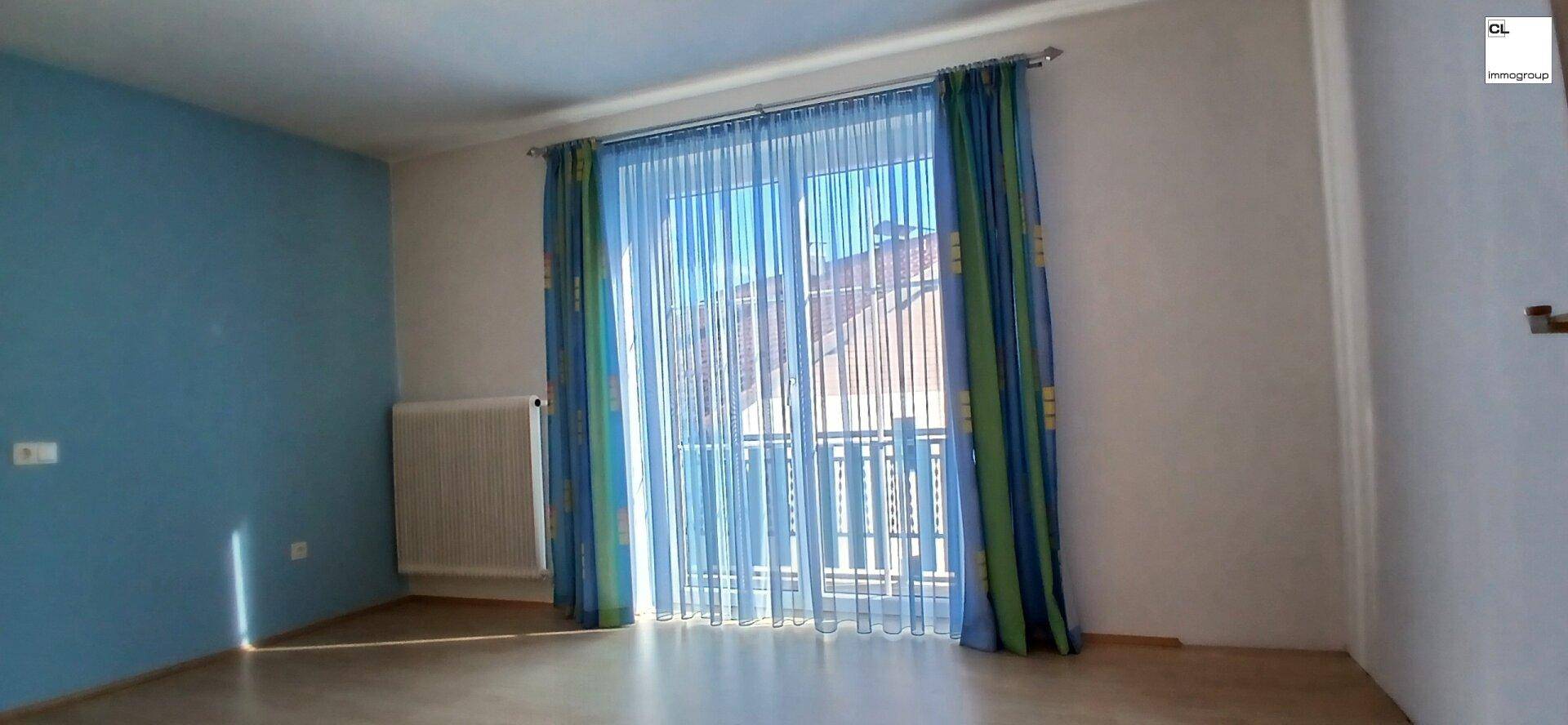 Heller Raum mit blauer Wand, großem Fenster und farbigen Vorhängen, ideal als Schlaf- oder Wohnzimmer.