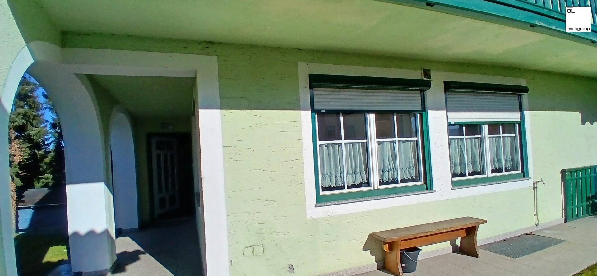 Überdachter Eingangsbereich im Erdgeschoss mit Fenstern, einer Holzbank und grüner Fassade.