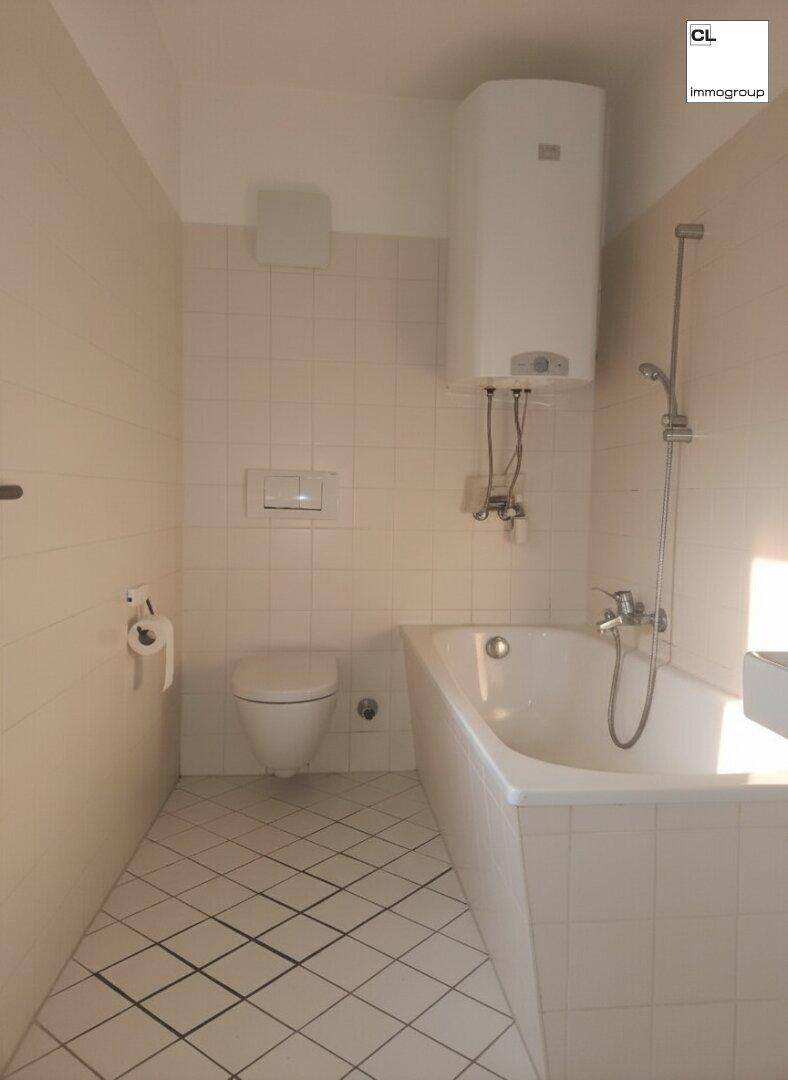 Badezimmer mit Badewanne, Toilette und Boiler, gefliest in hellem Beige mit diagonalen Fliesen.
