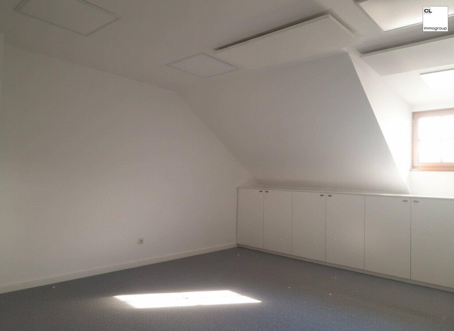 Dachgeschossbüro mit schrägen Wänden, Einbauschränken und Teppichboden, hell und geräumig.