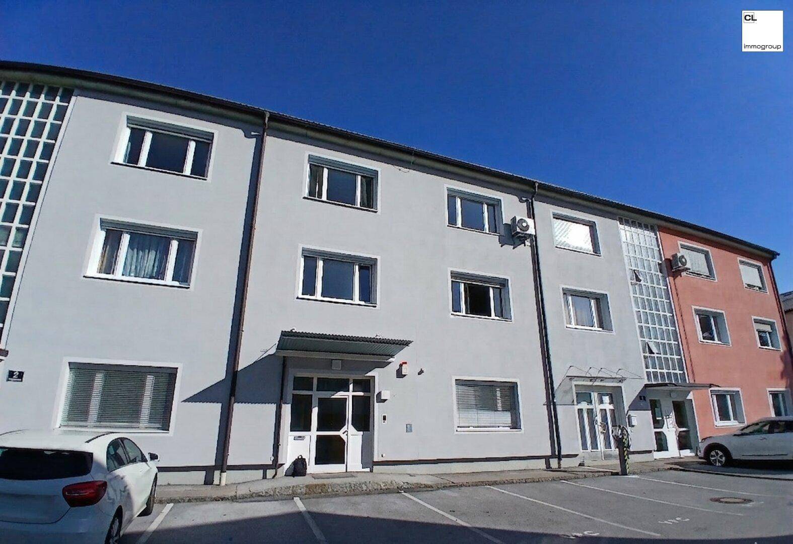 Mehrstöckiges Wohngebäude mit grauer und oranger Fassade, vielen Fenstern und Parkplätzen davor unter blauem Himmel.