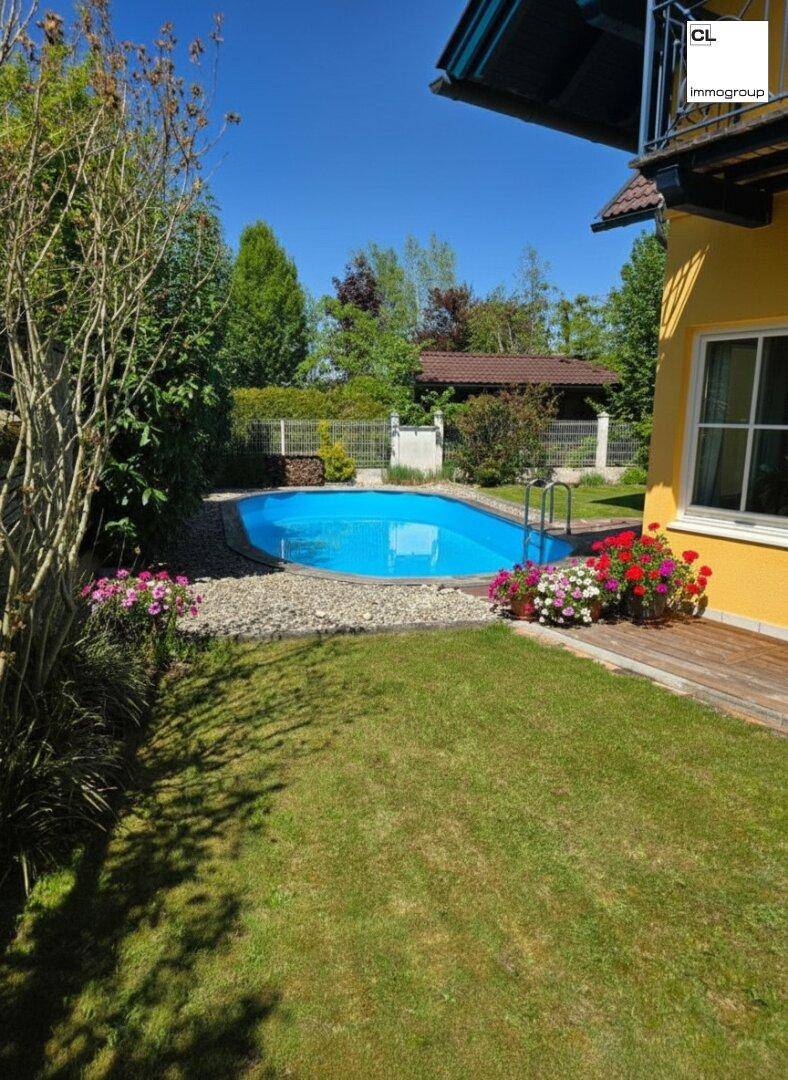 Ovaler Swimmingpool im Garten, umgeben von Blumenbeeten und Rasenfläche, unter blauem Himmel.