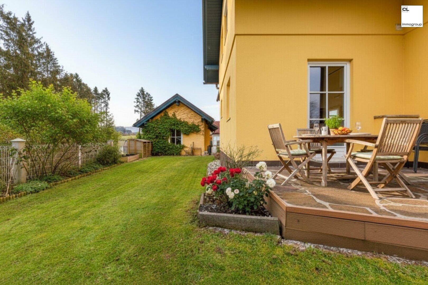 Steinplatten-Terrasse mit Gartenmöbeln und Blick auf den weitläufigen Rasen und ein Nebengebäude.