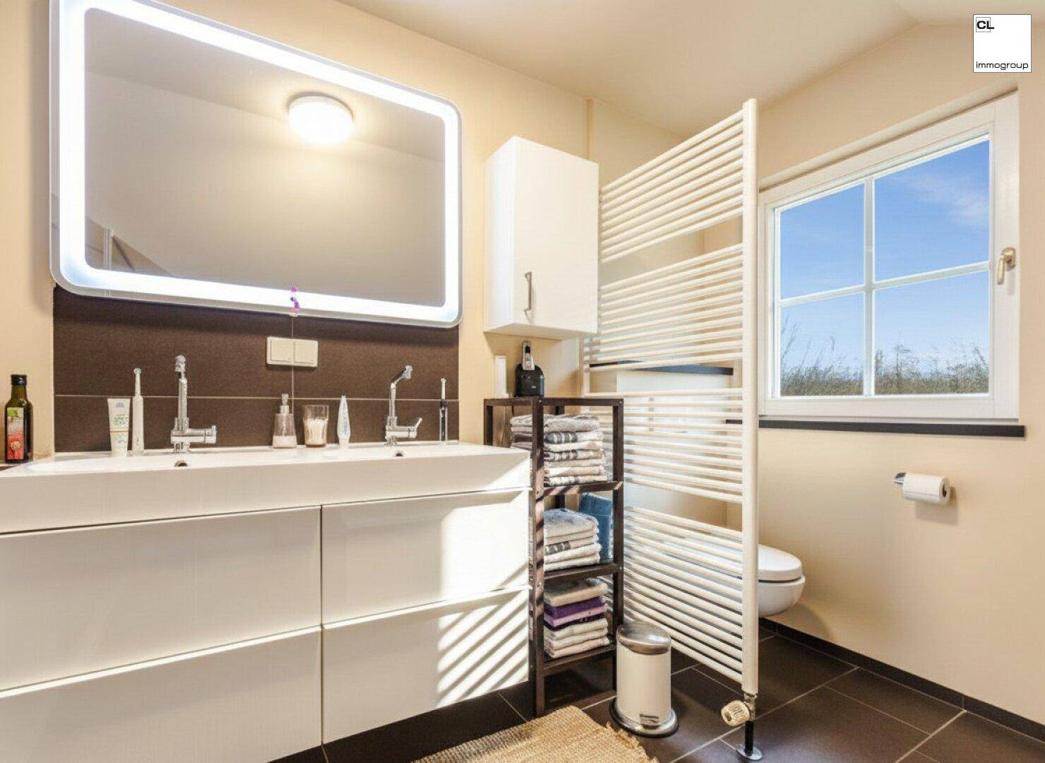 Modernes Badezimmer mit Doppelwaschbecken, beleuchtetem Spiegel, beheiztem Handtuchhalter und Fenster.