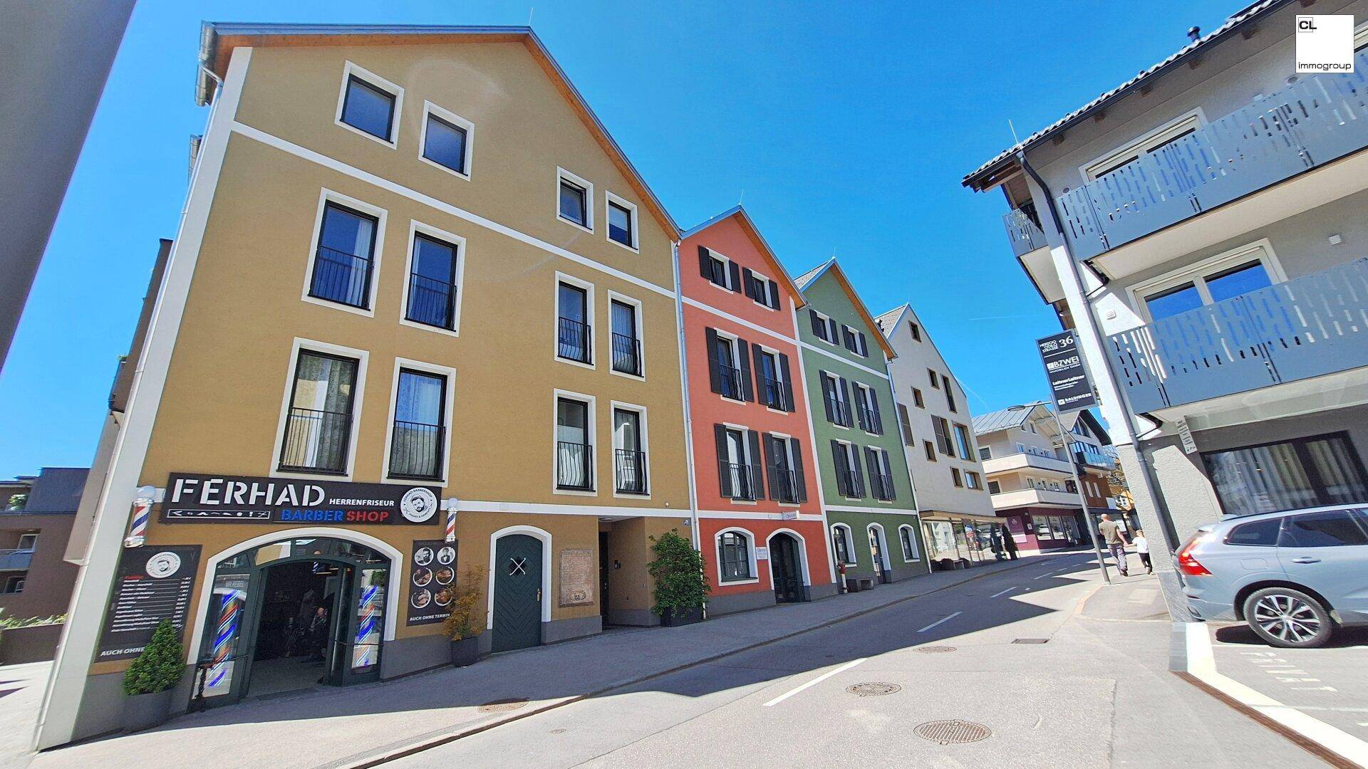Bunte Fassaden von Mehrfamilienhäusern entlang einer sonnigen Straße mit Geschäften im Erdgeschoss.