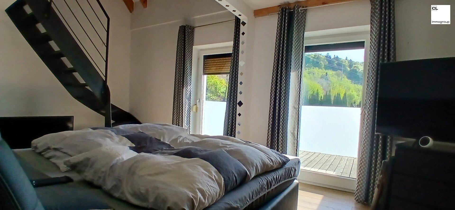 Gemütliches Schlafzimmer mit Doppelbett, großen Fenstern und Zugang zu einer Treppe.