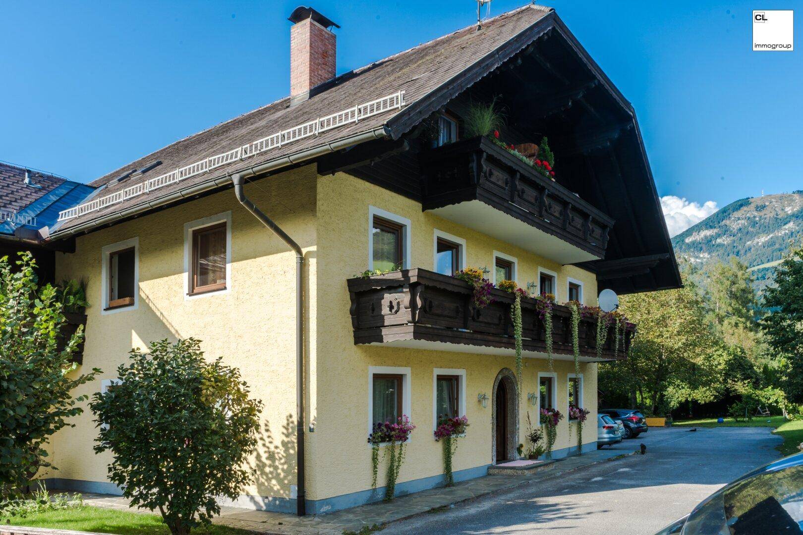 Traditionelles gelbes Gebäude mit Holzbalkonen, Blumenkästen und Bergblick unter strahlend blauem Himmel.