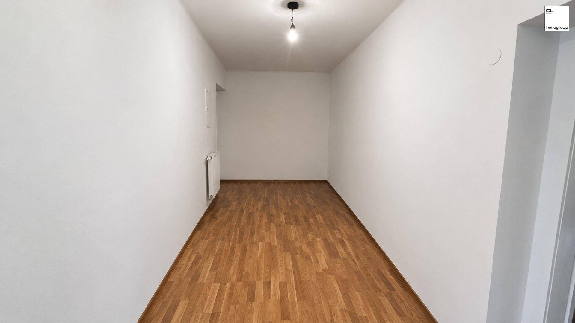 Langer, schmaler Raum mit hellen Wänden und Holzfußboden, beleuchtet von einer einzelnen Deckenlampe.