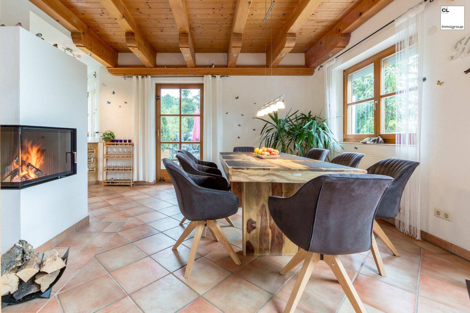 Essbereich mit rustikalem Holztisch, Kamin und großen Fenstern mit Blick ins Grüne.