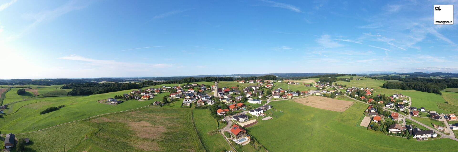 Weitläufige Panorama-Luftaufnahme eines ländlichen Dorfes, eingebettet in grüne Felder und Natur.