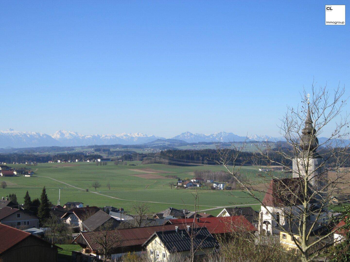 Blick über ein Dorf mit Kirchturm auf weite Felder und schneebedeckte Berge in der Ferne.