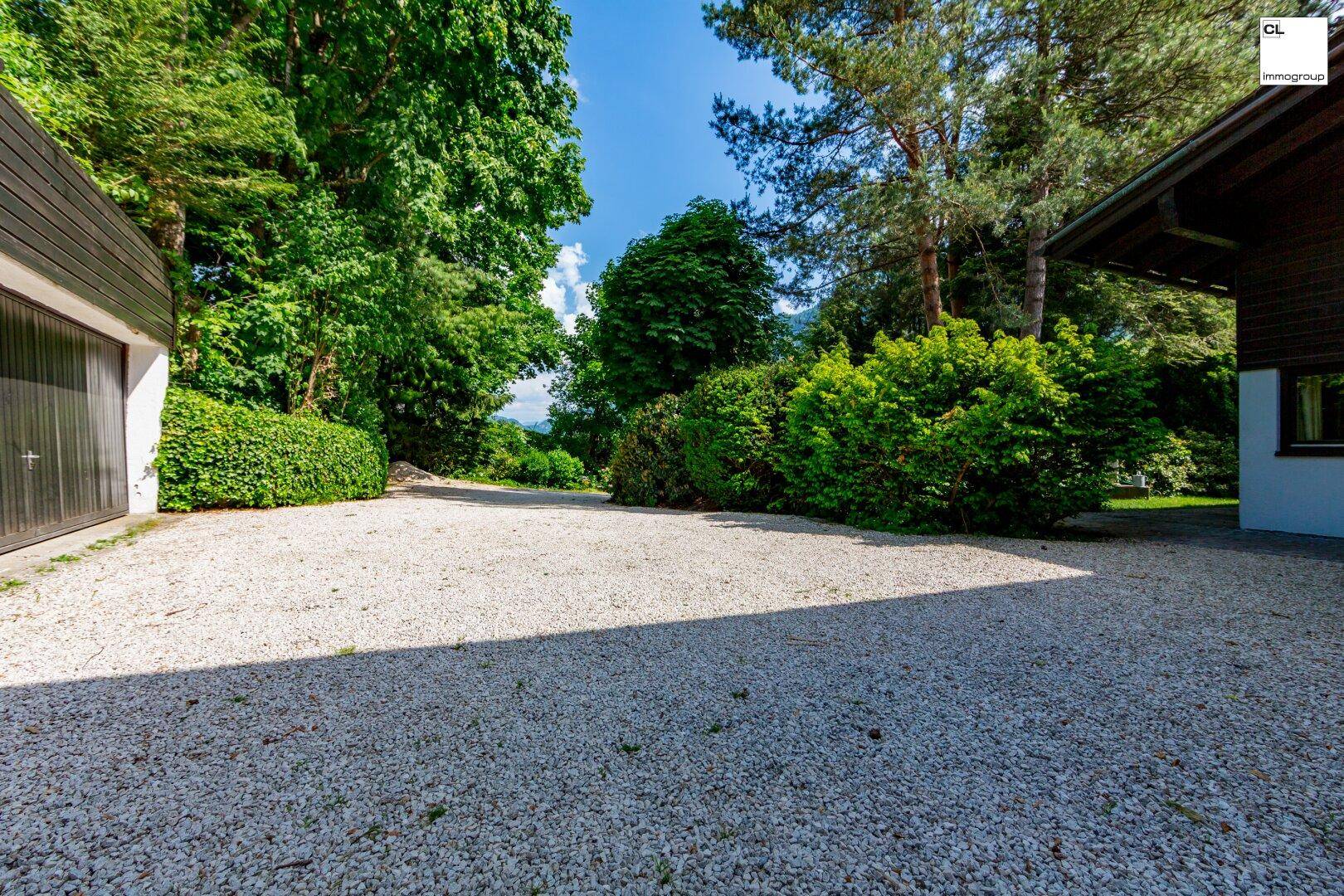 Geräumige Kiesauffahrt führt zu einer Garage, umgeben von dichtem Grün und hohen Bäumen.
