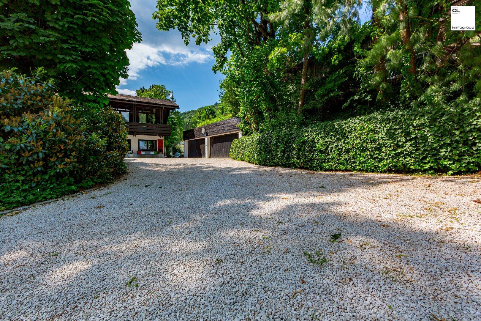 Breite Kiesauffahrt zum Haus und zur Garage, eingebettet in eine grüne und bewachsene Umgebung.