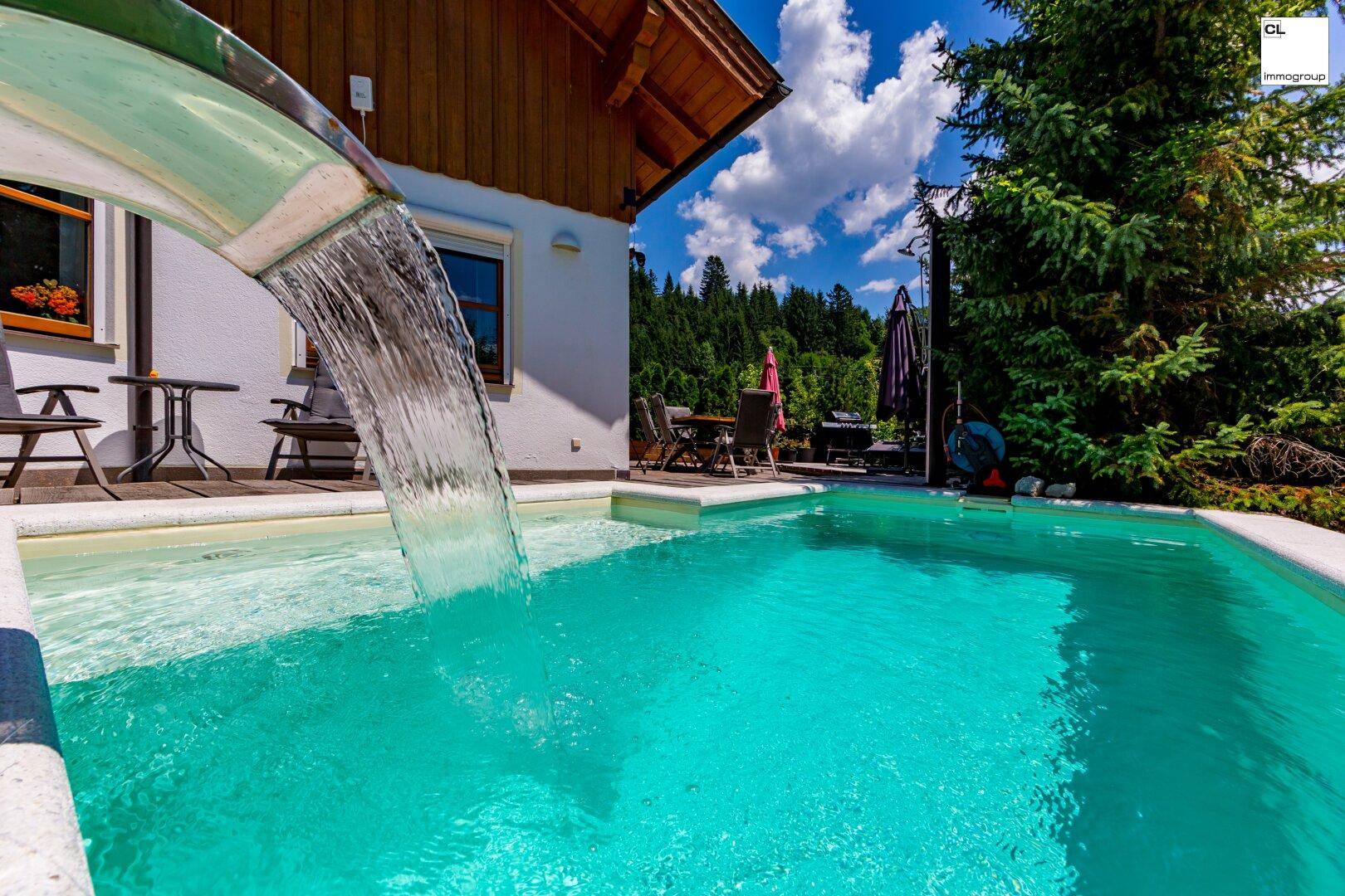 Außenpool mit Wasserspiel und Holzdeck, umgeben von grünen Bäumen unter blauem Himmel.