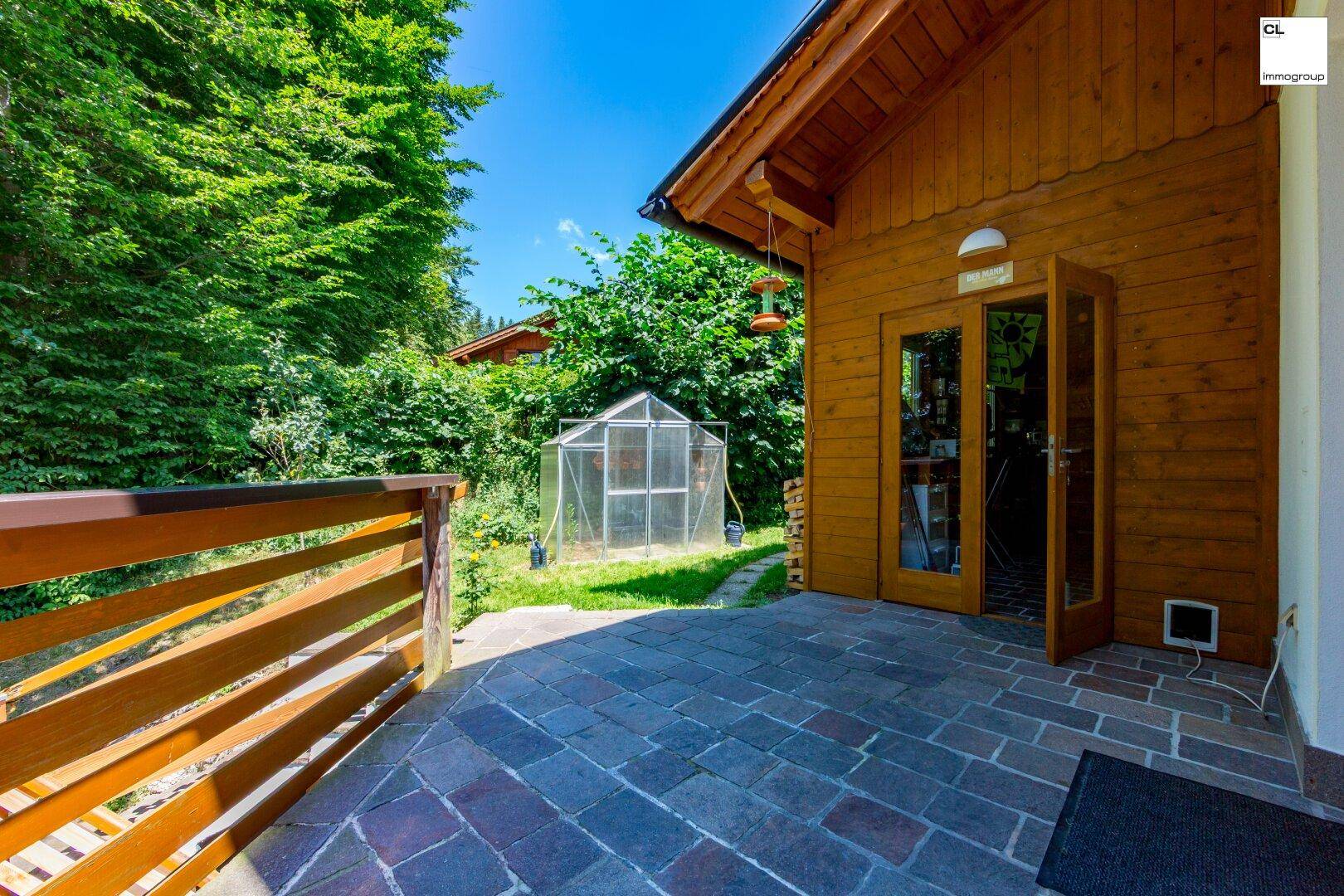 Gepflasterter Bereich im Garten mit Gewächshaus und Zugang zu einem Nebengebäude.