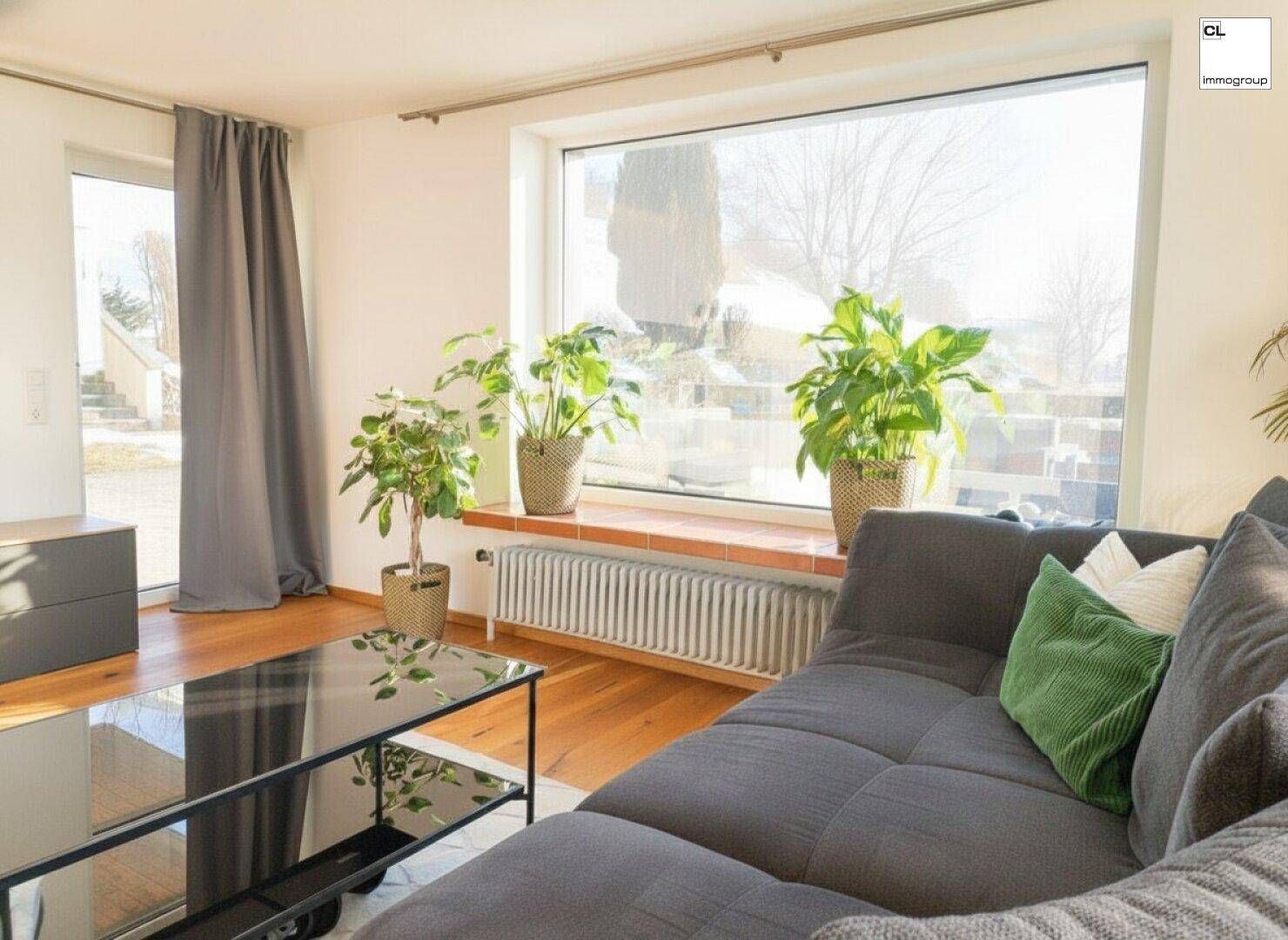 Helles Wohnzimmer mit großem Sofa, Holzboden und großen Fenstern, die viel Tageslicht hereinlassen.