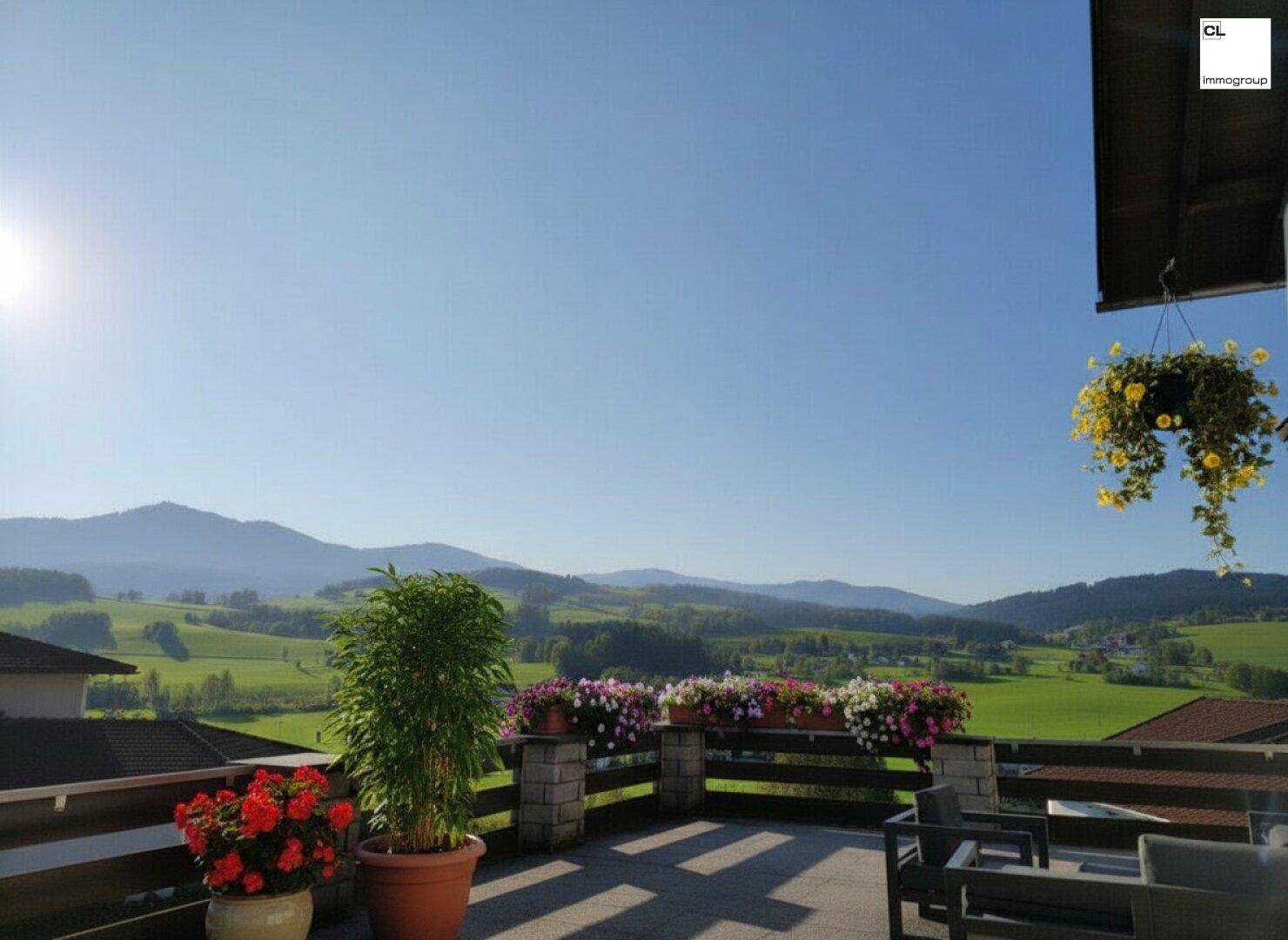 Sonniger Terrassenbereich mit Blumenkästen und Sitzgelegenheiten, umgeben von grünen Hügeln und Bergen.