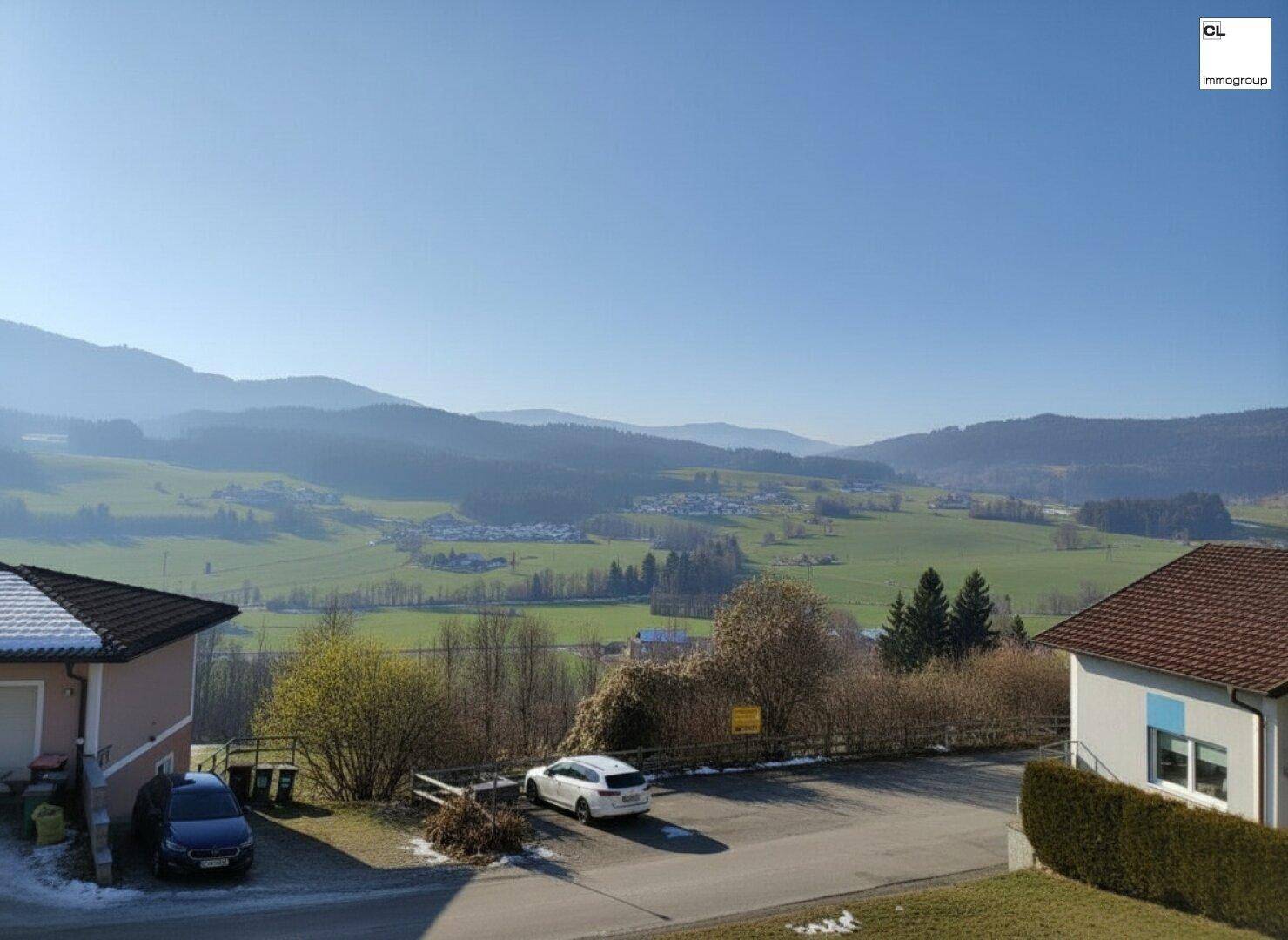 Weitblick über die ländliche Umgebung mit Häusern, einer Straße und grünen Hügeln unter blauem Himmel.