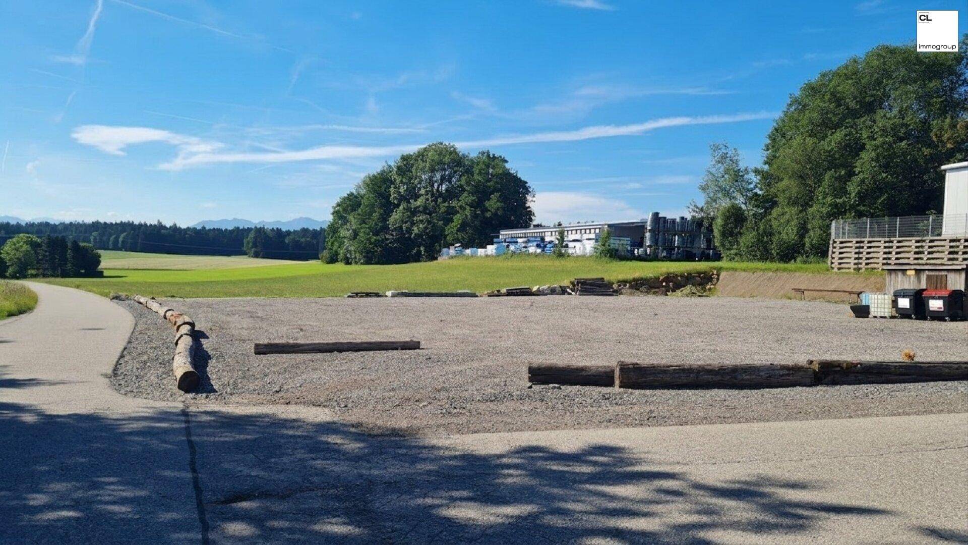 Weitläufiger Schotterparkplatz neben einer Straße mit grünen Feldern und Bäumen im Hintergrund unter blauem Himmel.
