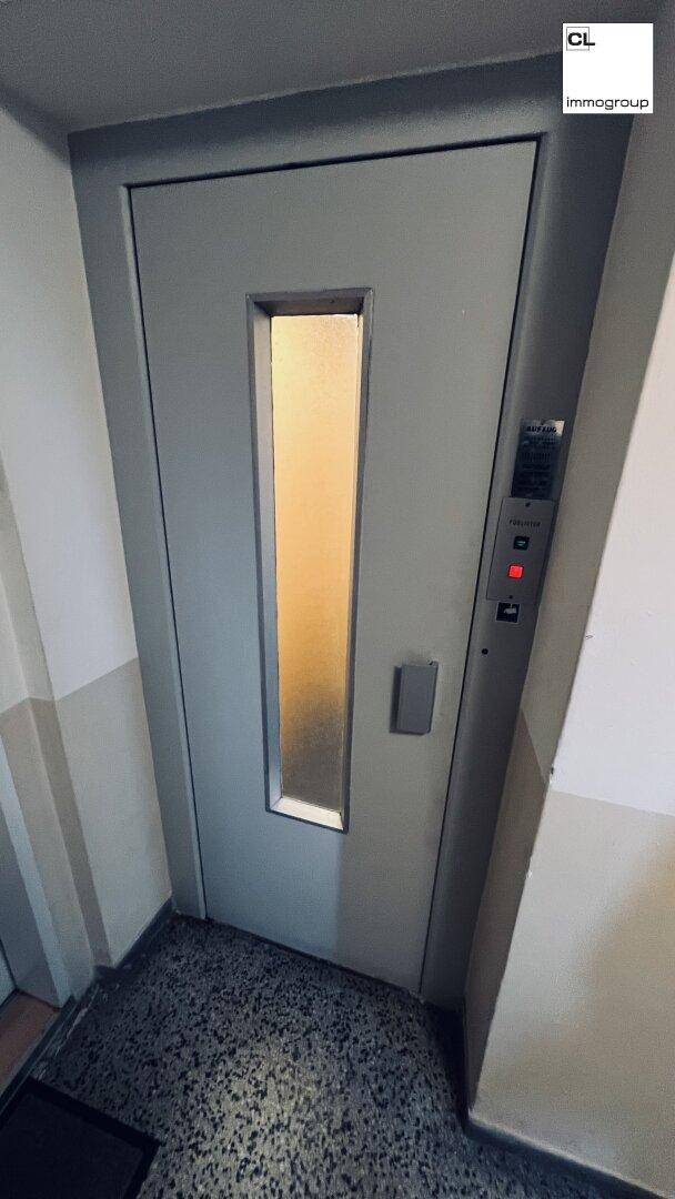 Graue Aufzugstür mit vertikalem Milchglasfenster in einem Flur, bereit zur Nutzung.