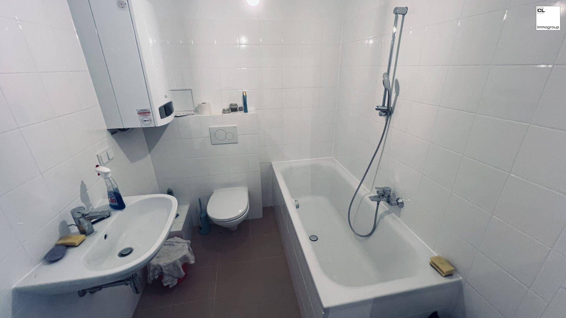 Helles Badezimmer mit weißen Fliesen, ausgestattet mit Waschbecken, Toilette und einer Badewanne mit Duschfunktion.
