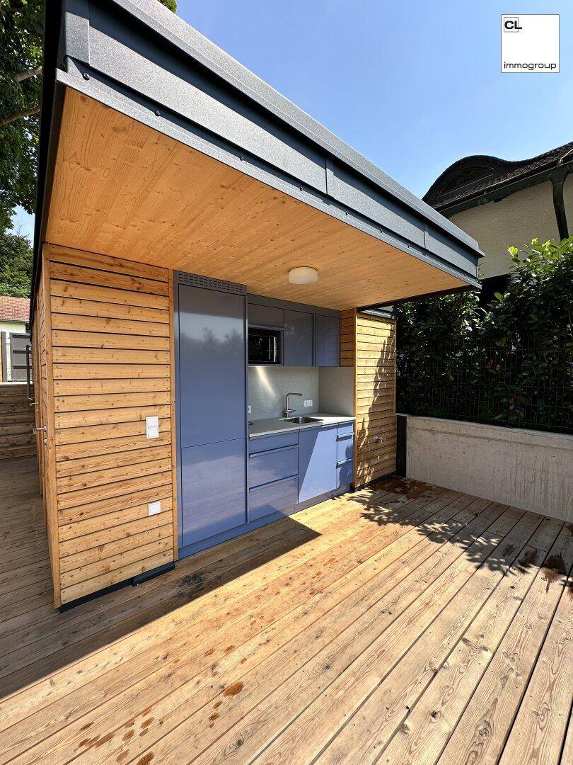 Moderne Außenküche mit blauen Schränken und Holzwänden auf einer sonnigen Holzterrasse.