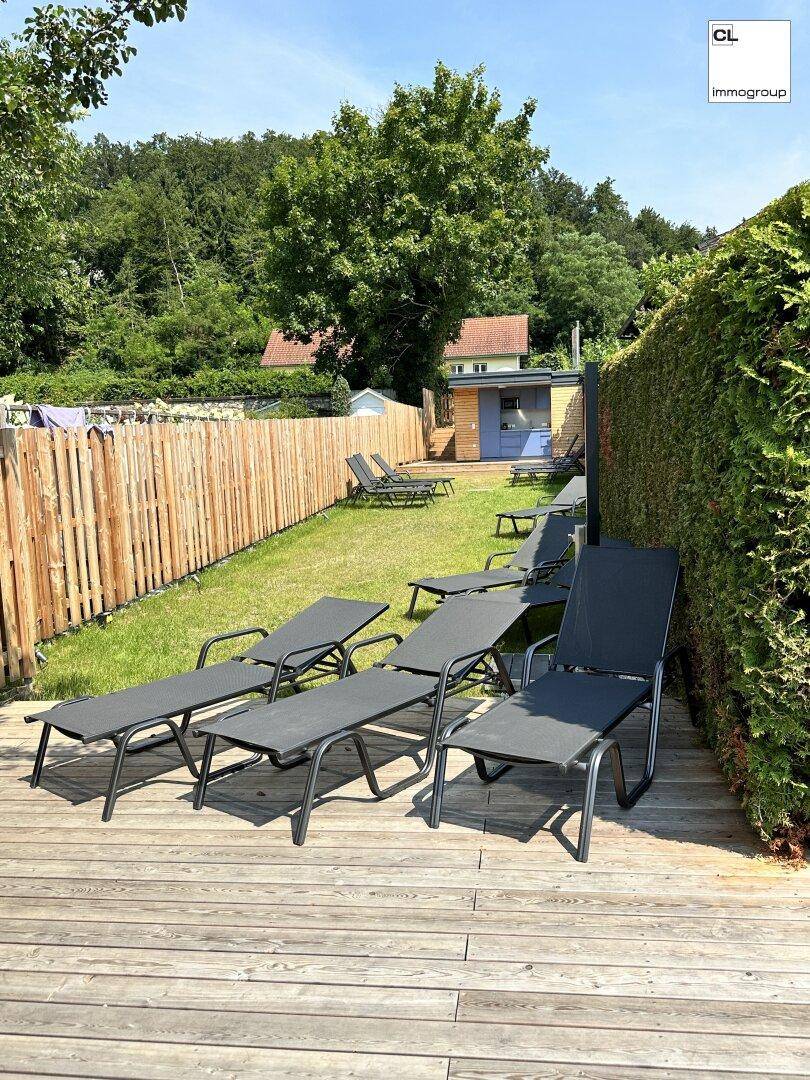 Entspannungsbereich im Garten mit mehreren Sonnenliegen auf einer Holzterrasse und grüner Hecke.