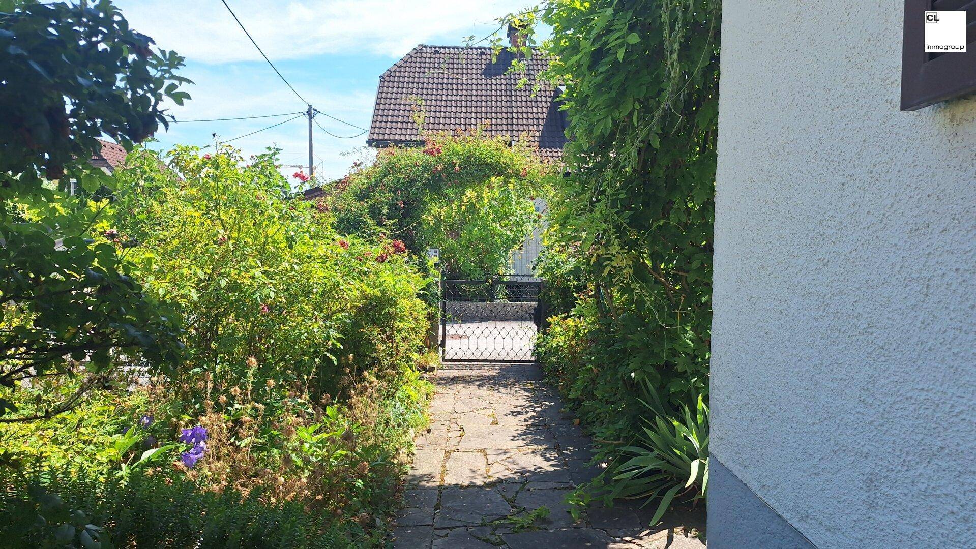 Gepflasterter Gartenweg führt durch dichte Hecken zu einem schmiedeeisernen Gartentor.