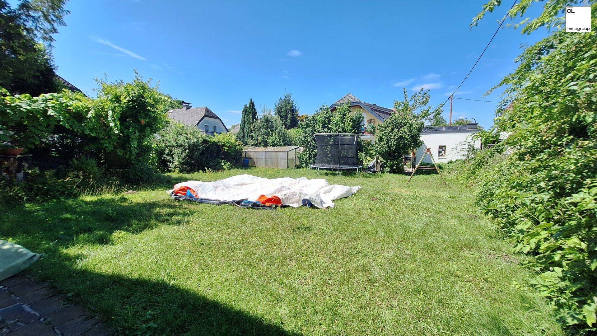 Großer, sonniger Garten mit Rasenfläche, Trampolin und kleinem Gewächshaus im Hintergrund.