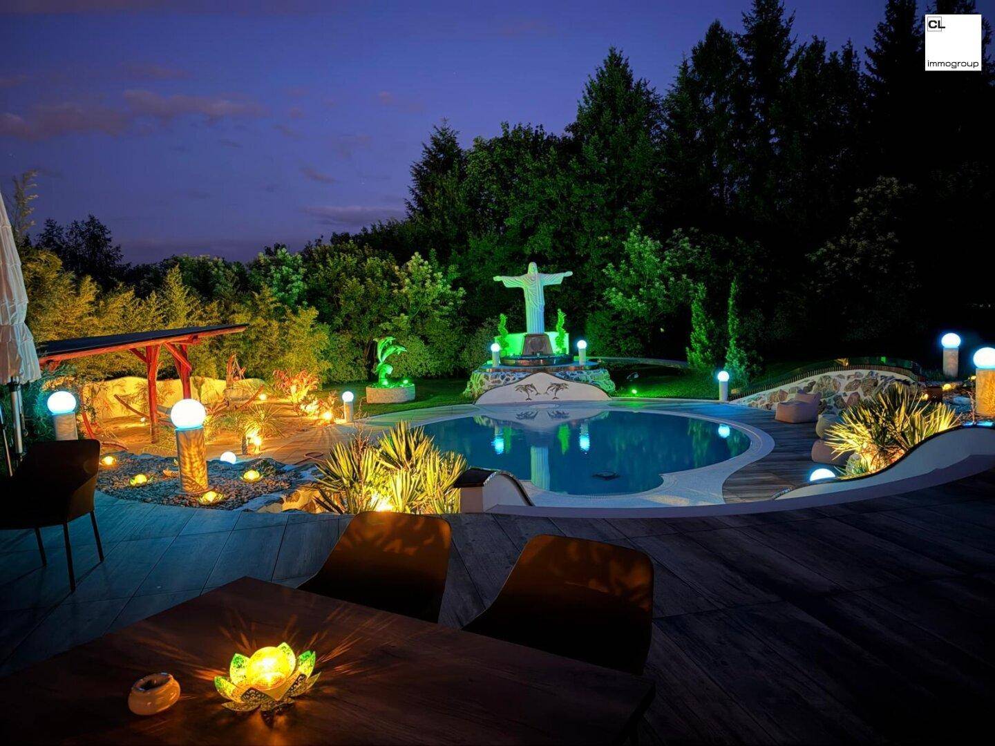 Beleuchteter Poolbereich im Garten bei Nacht mit Holzterrasse und dekorativer Statue.
