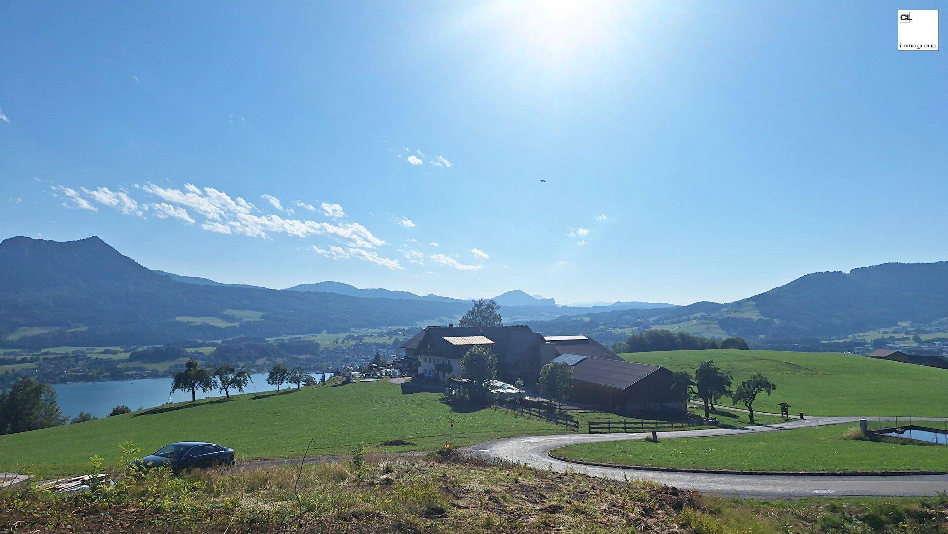 Weitläufiges Anwesen mit mehreren Gebäuden auf einem grünen Hügel, mit See- und Bergblick unter strahlendem Sonnenschein.