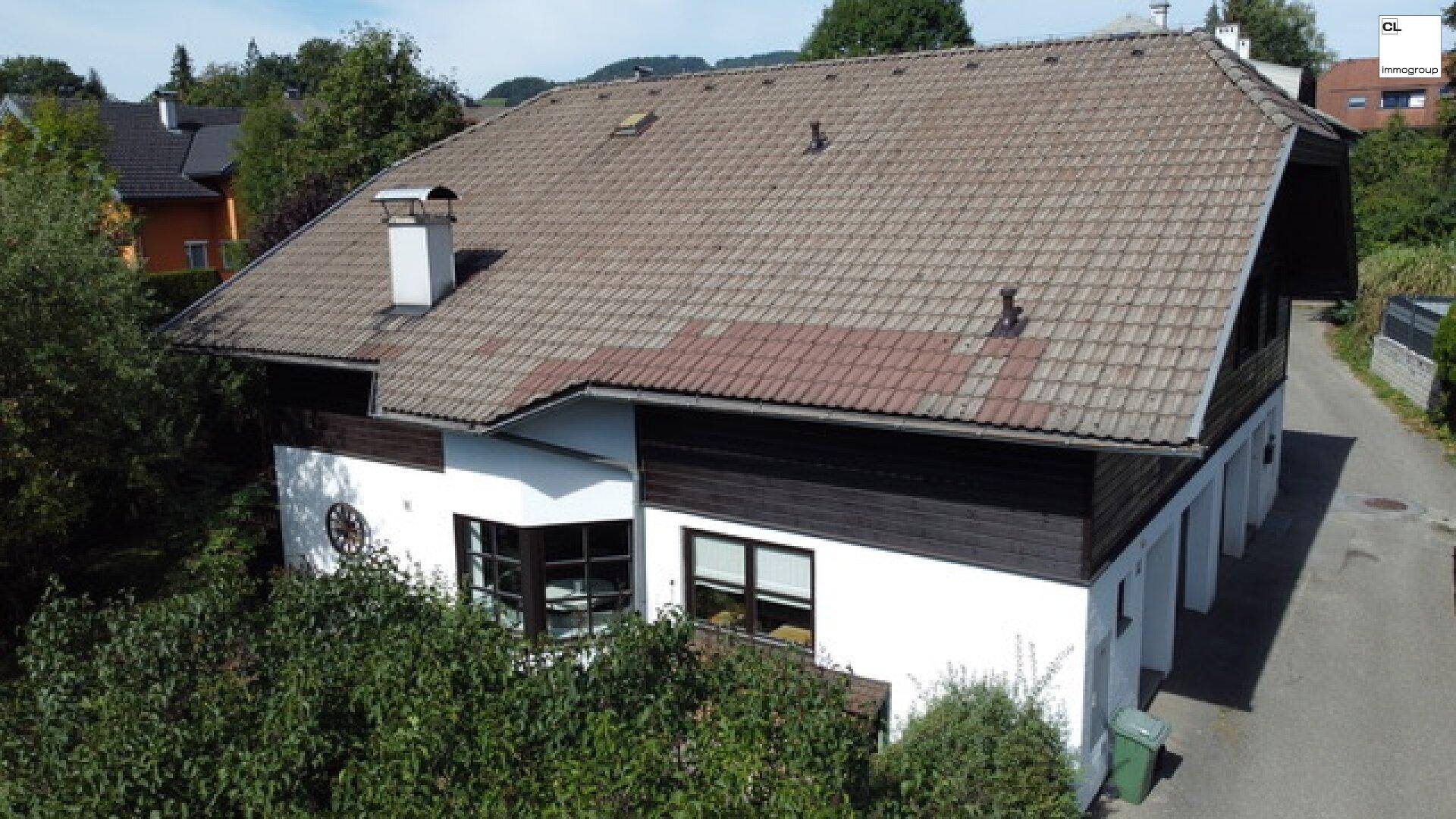 Haus mit weißer Fassade, dunklen Holzelementen und einem Erker, umgeben von üppiger Vegetation.