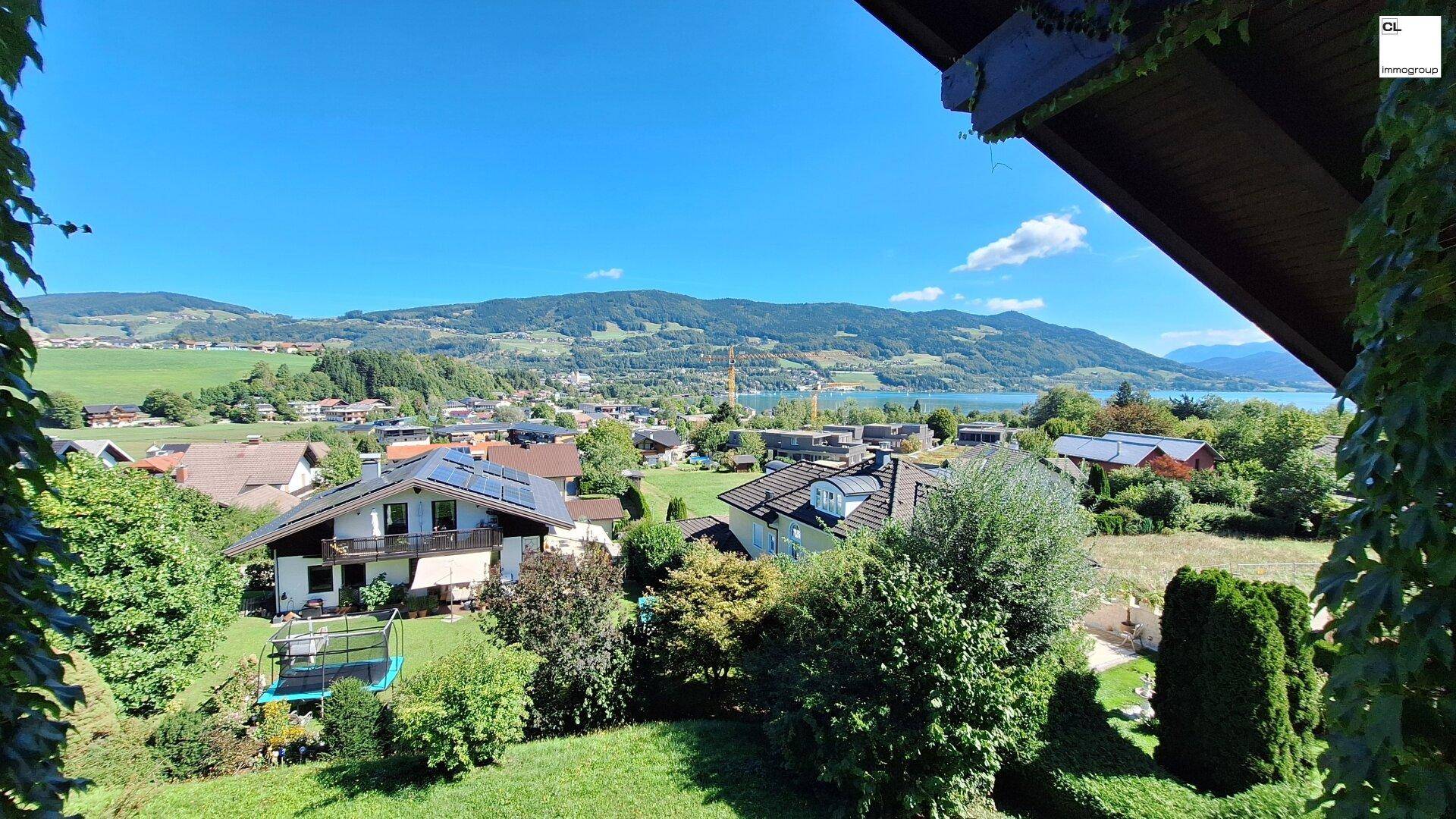 Weitläufiger Blick über die Landschaft mit Häusern, viel Grün und einem See im Hintergrund an einem sonnigen Tag.