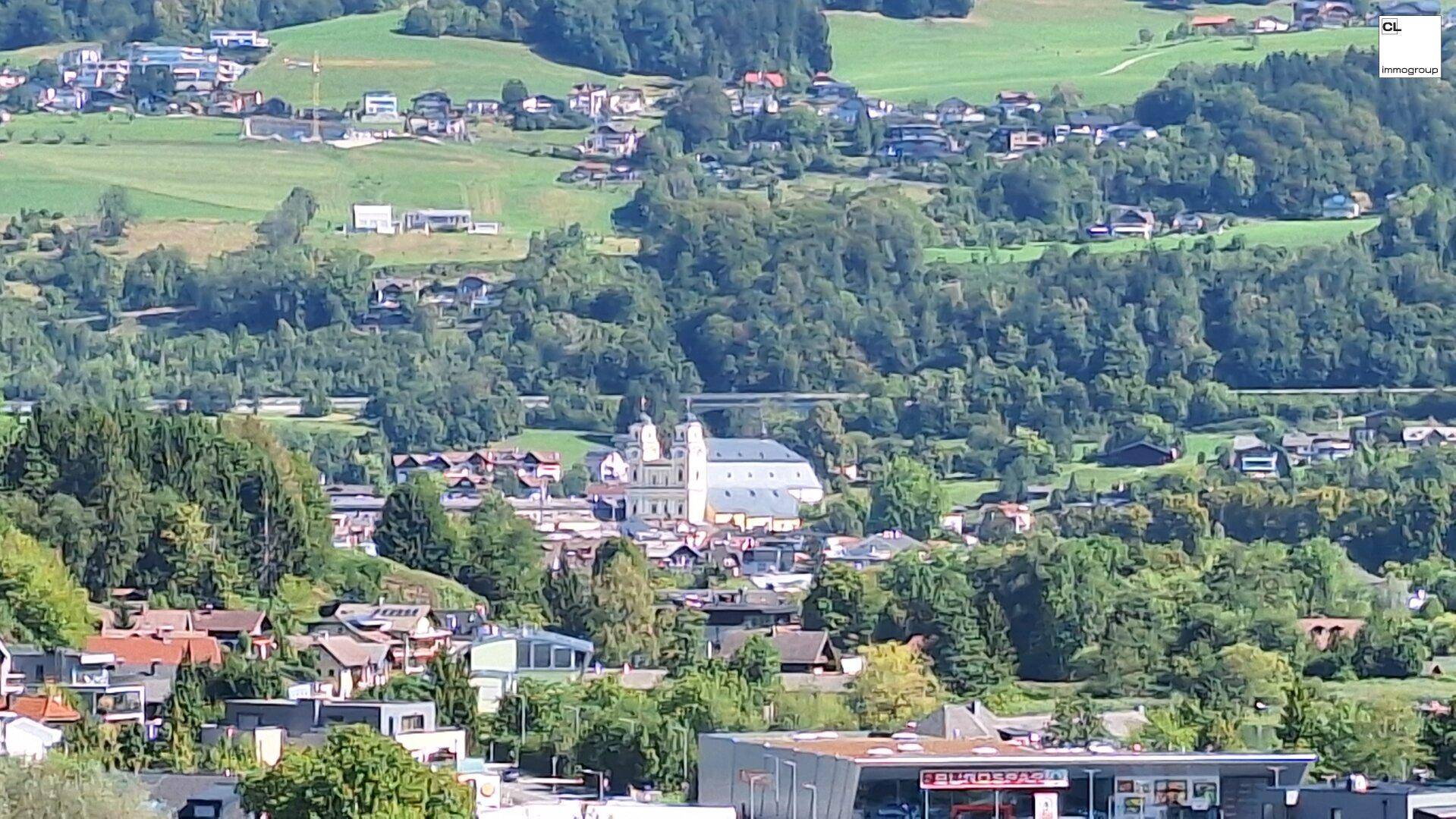 Luftaufnahme einer Stadt mit einer großen Kirche, umgeben von grünen Hügeln und Häusern unter blauem Himmel.