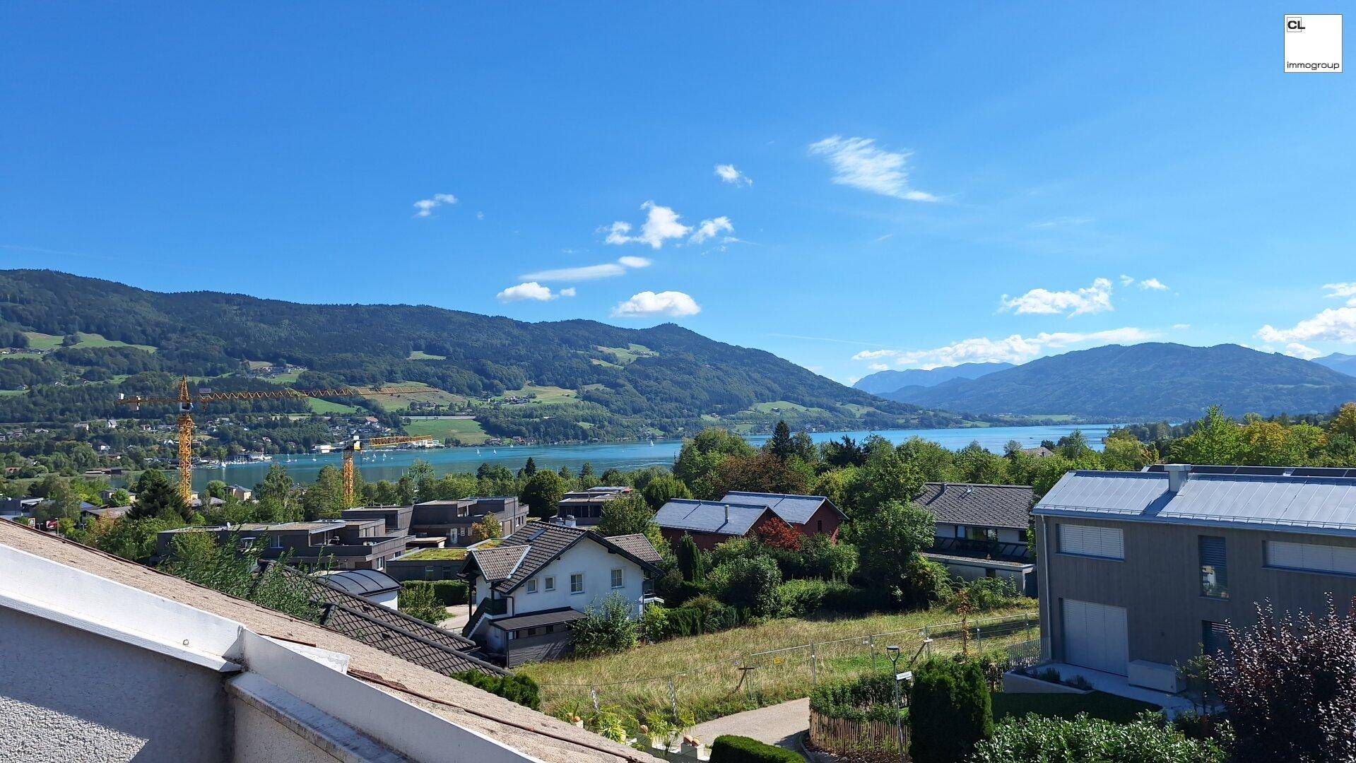Malerischer Ausblick auf den See und die Berge unter blauem Himmel mit einigen Wolken und Dächern im Vordergrund.
