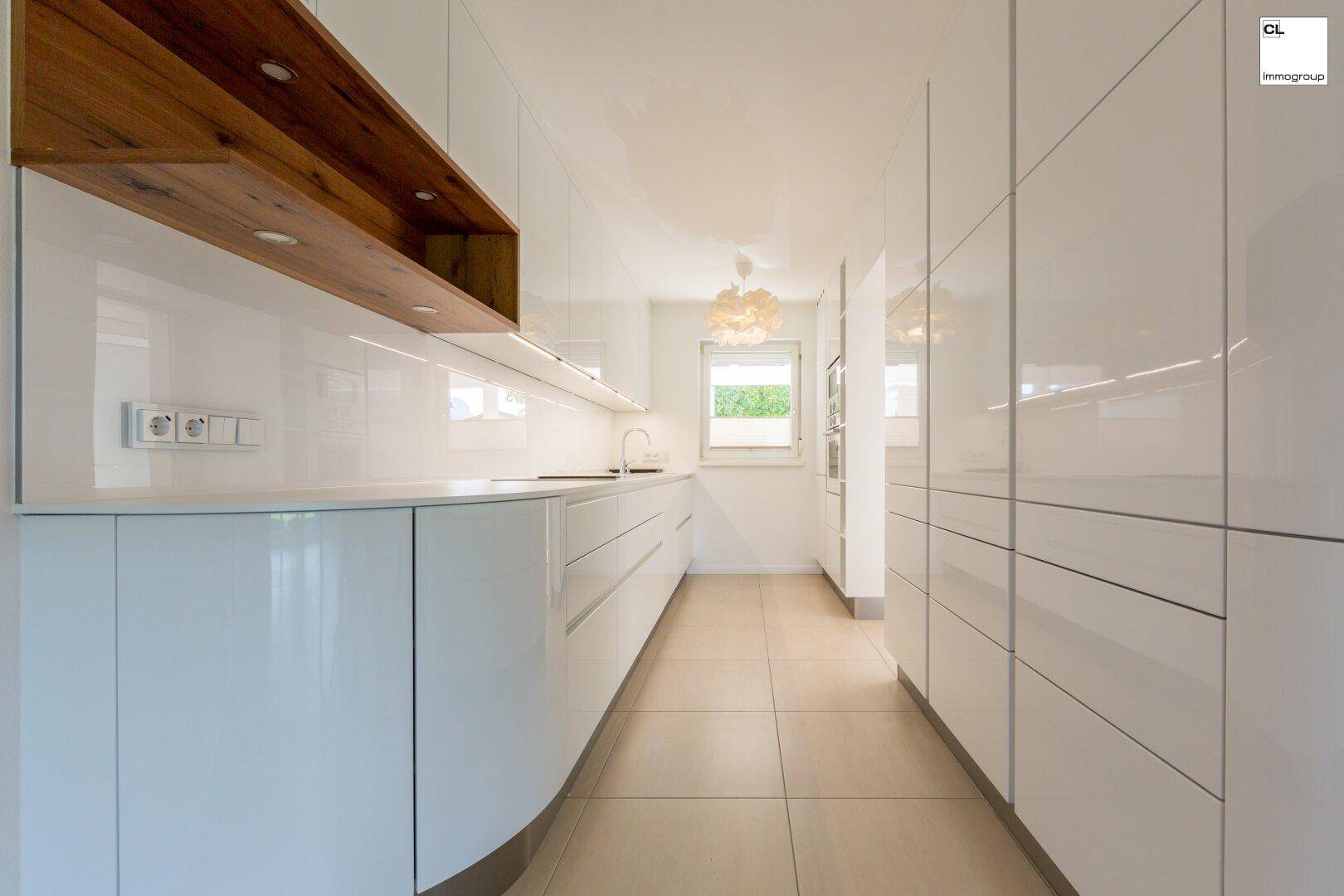 Moderne, helle Küche mit weißen Hochglanzfronten, Holzakzenten und Fenster.