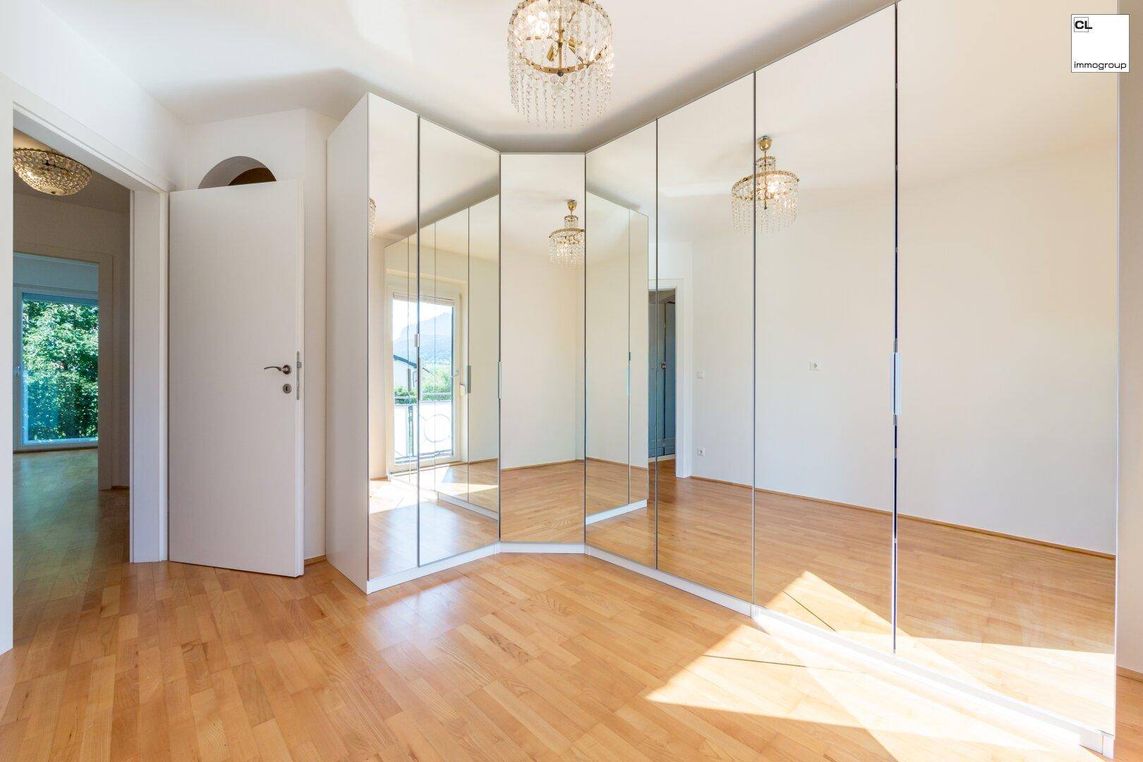 Schlafzimmer mit Spiegelschränken, Parkettboden und Zugang zu einem sonnigen Balkon.