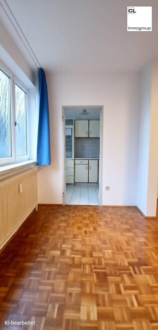Ein leerer Raum mit Parkettboden und Blick durch eine Tür in eine angrenzende Küche.