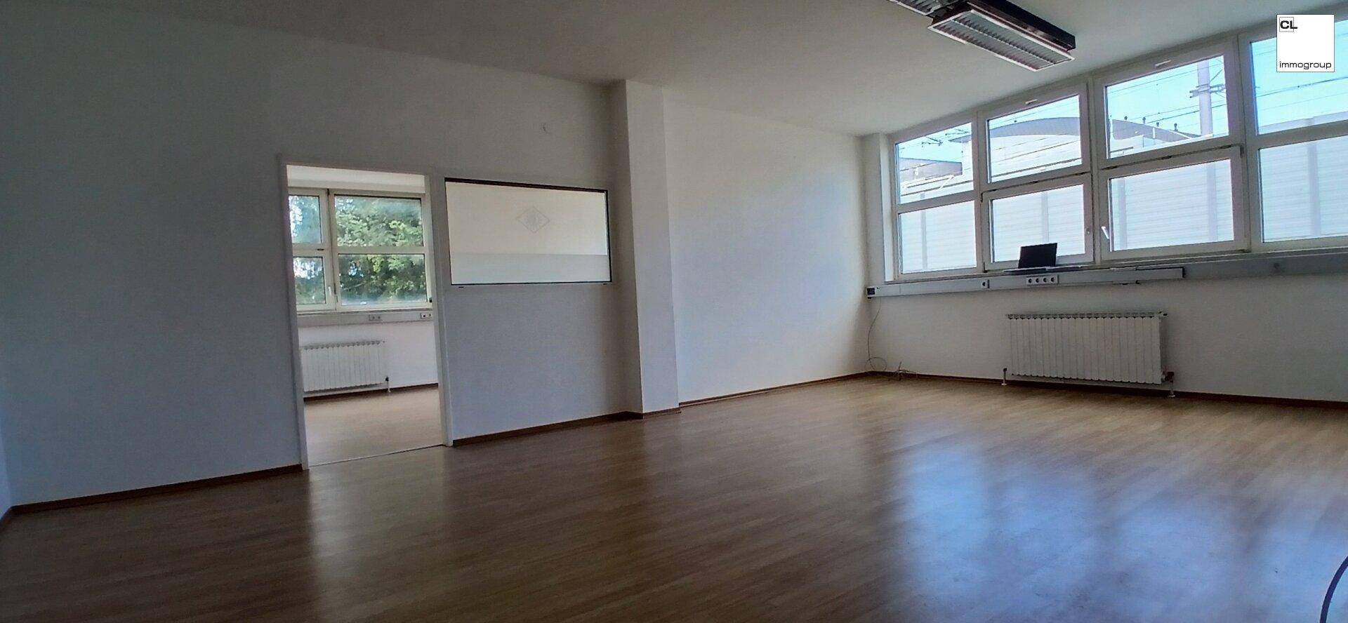 Heller Büroraum mit großen Fenstern, Holzboden und Verbindung zu einem Nebenraum, bietet viel Tageslicht.
