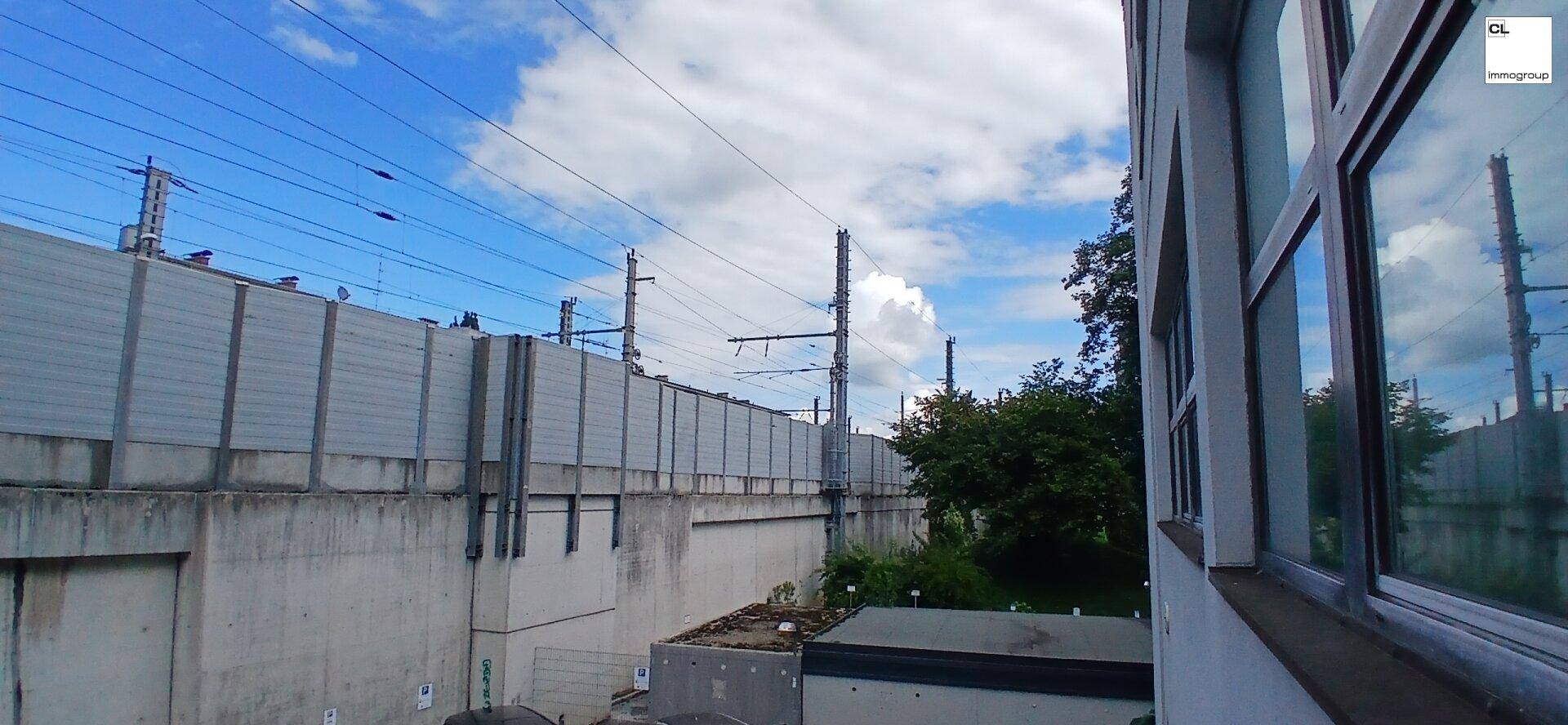 Blick aus dem Fenster auf Bahngleise mit Oberleitung, Lärmschutzwand und etwas Grünfläche.